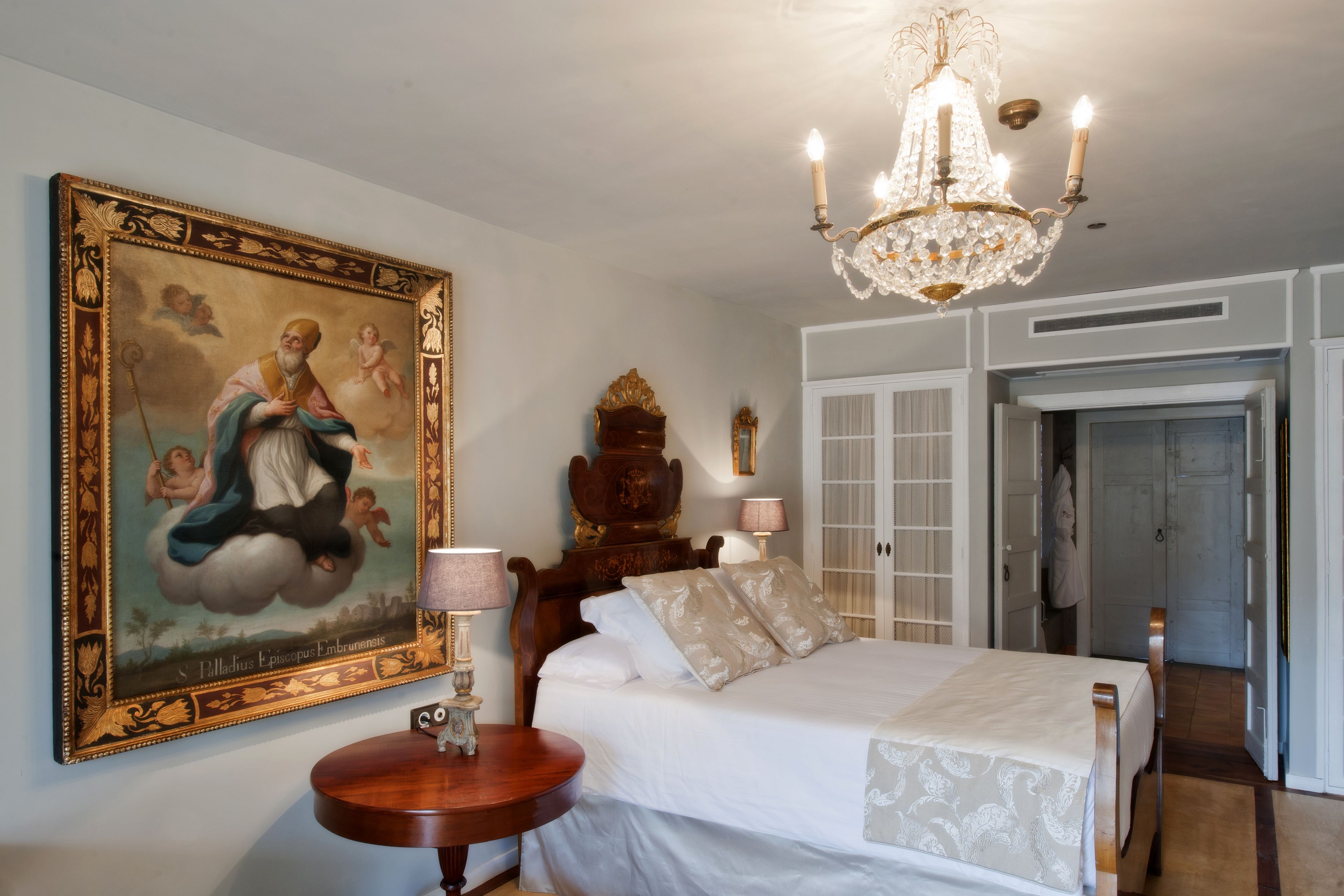 Un dormitorio con un cuadro grande y ornamentado, una araña de cristal, una cama de madera y una mesita de noche.
