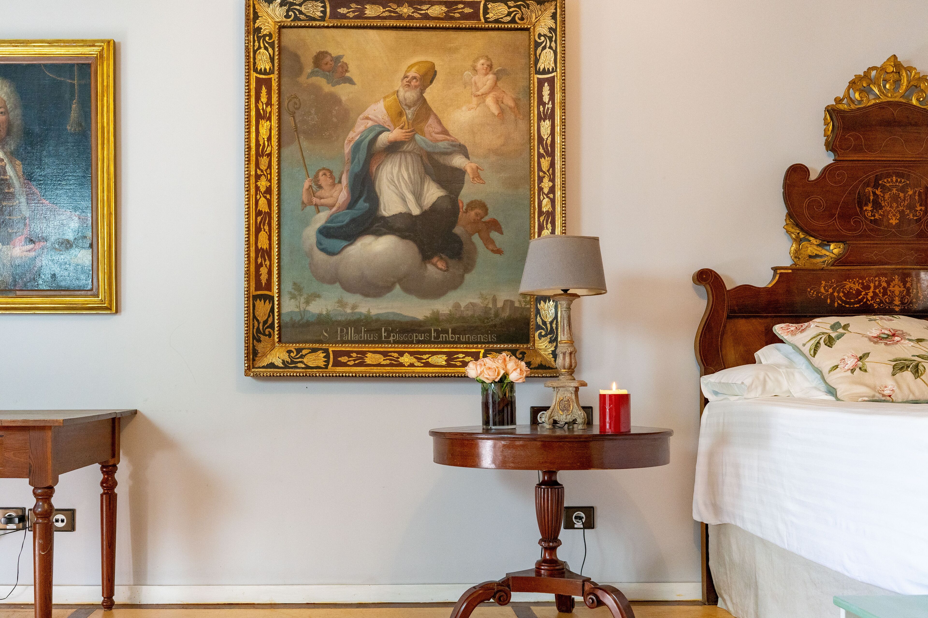 A bedroom with a bed, a nightstand, a table with a lamp, and a wall painting.