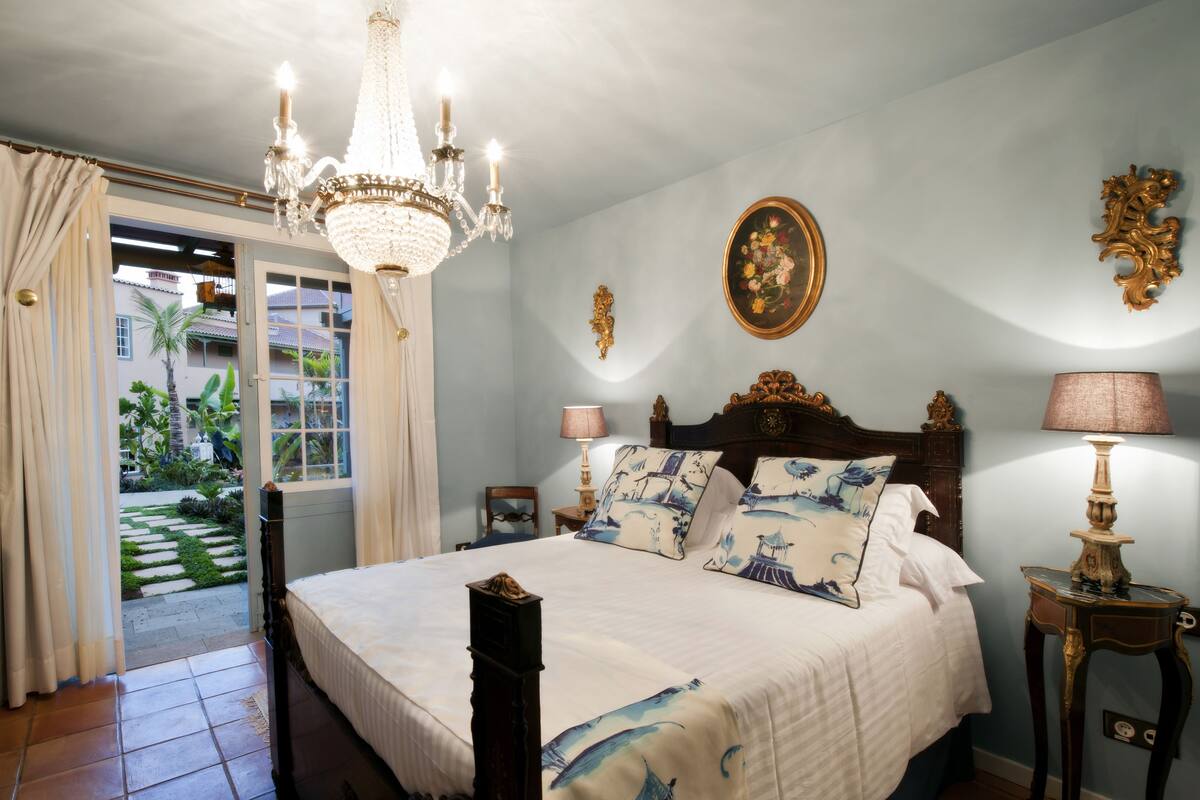 A bedroom with a large bed, a chandelier, a bedside table with a lamp, and a view of the outside.
