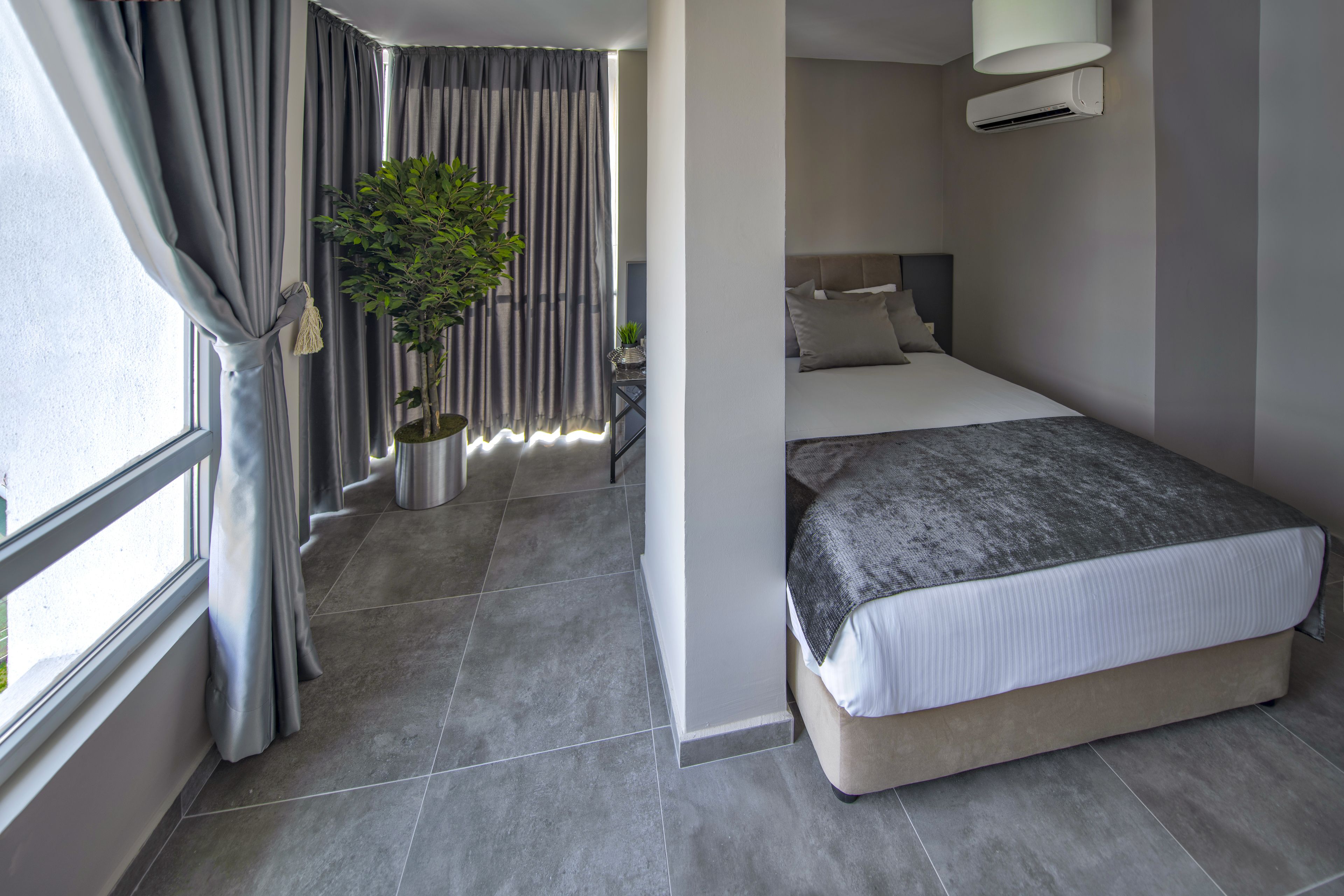 Une chambre d’hôtel moderne avec un grand lit, des rideaux gris, une plante en pot et une fenêtre donnant sur un beau paysage.