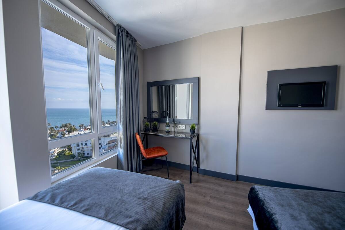 A hotel room with a large window, a desk with a chair, a bed, and a flat-screen TV mounted on the wall.