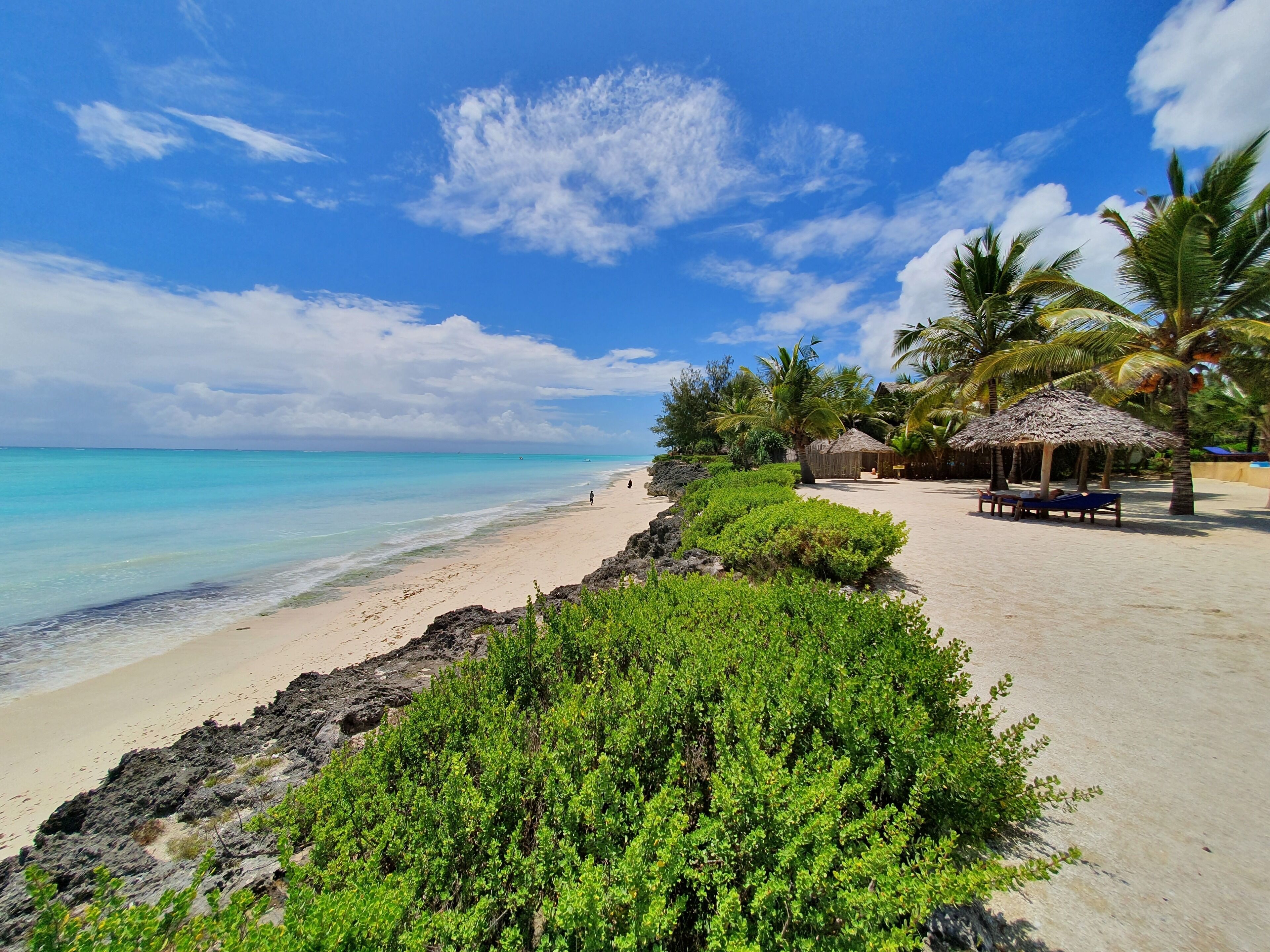 Ein Strand mit klarem türkisblauem Wasser, einem sandigen Ufer und üppiger Vegetation. Es sind Palmen und ein Gebäude mit einem Reetdach zu sehen.