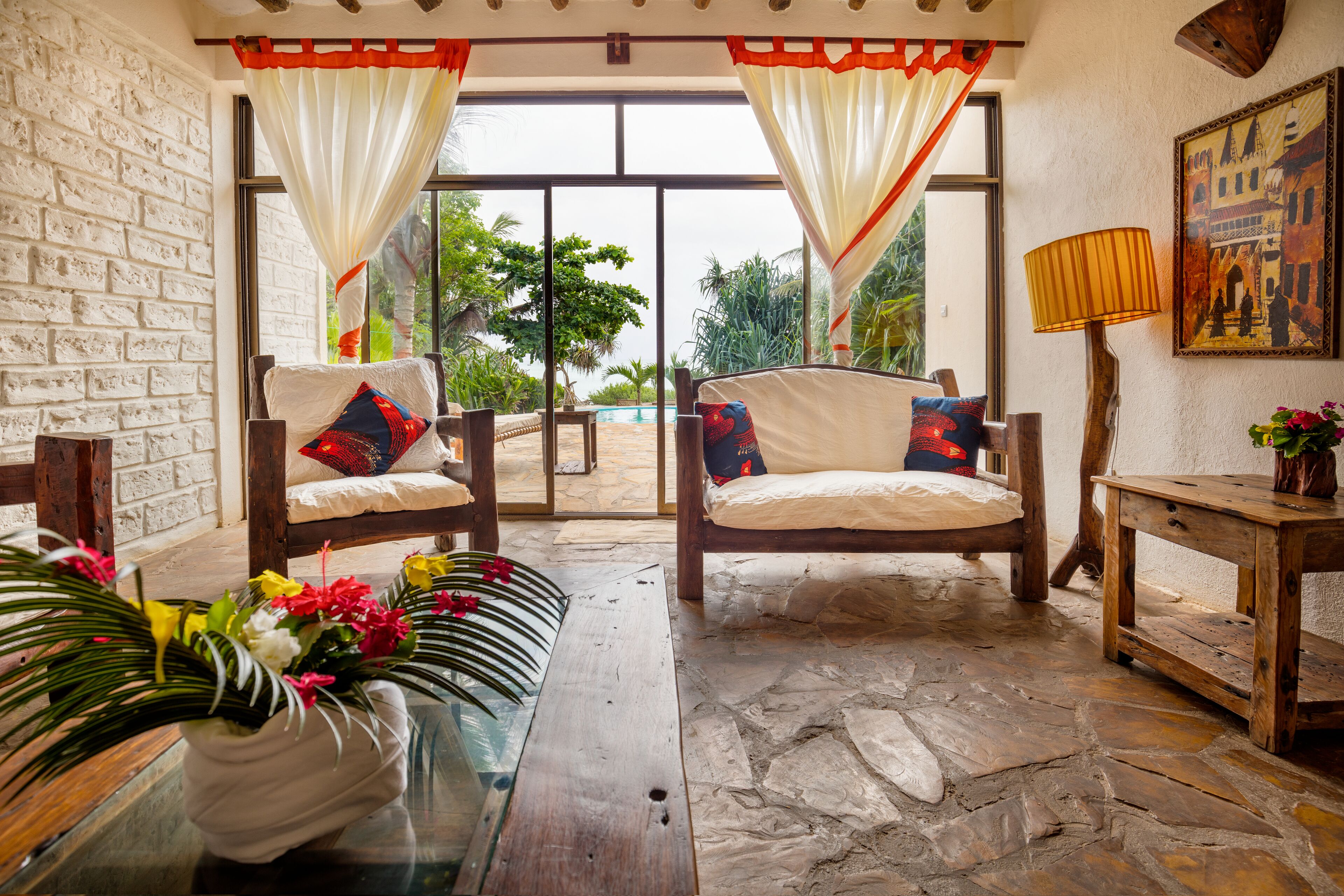 Una habitación con suelo de piedra, dos sillas de madera con cojines, una mesa de cristal con un jarrón de flores, una mesita de noche de madera con una lámpara, y un ventanal con cortinas.