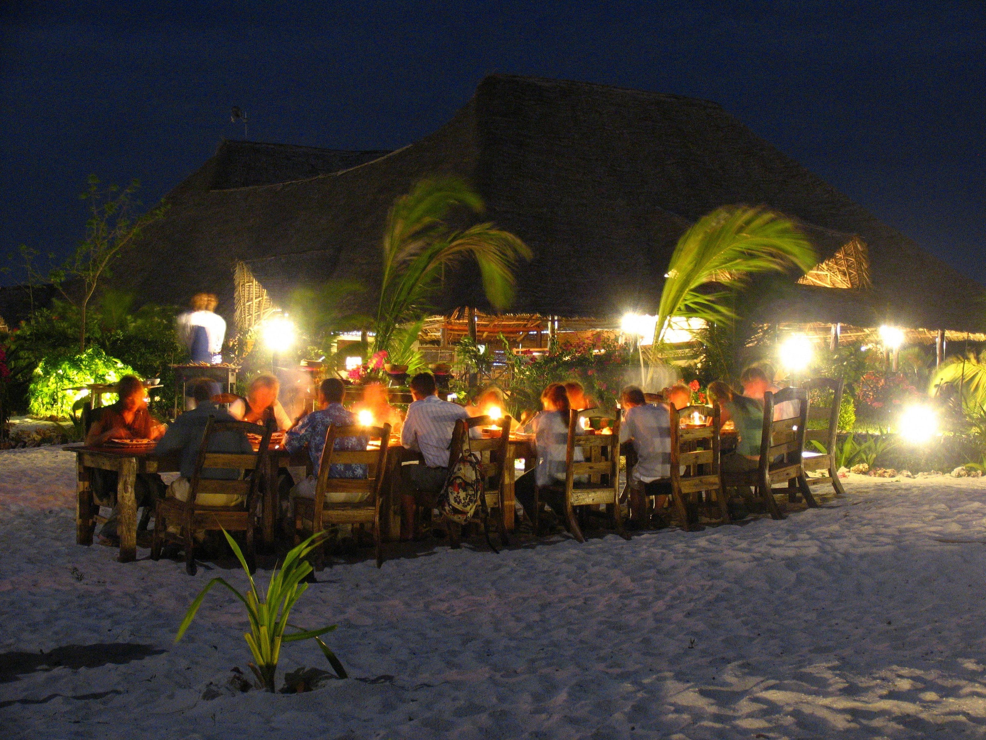 Un grupo de personas cenando en un restaurante al aire libre con techos de paja y palmeras.