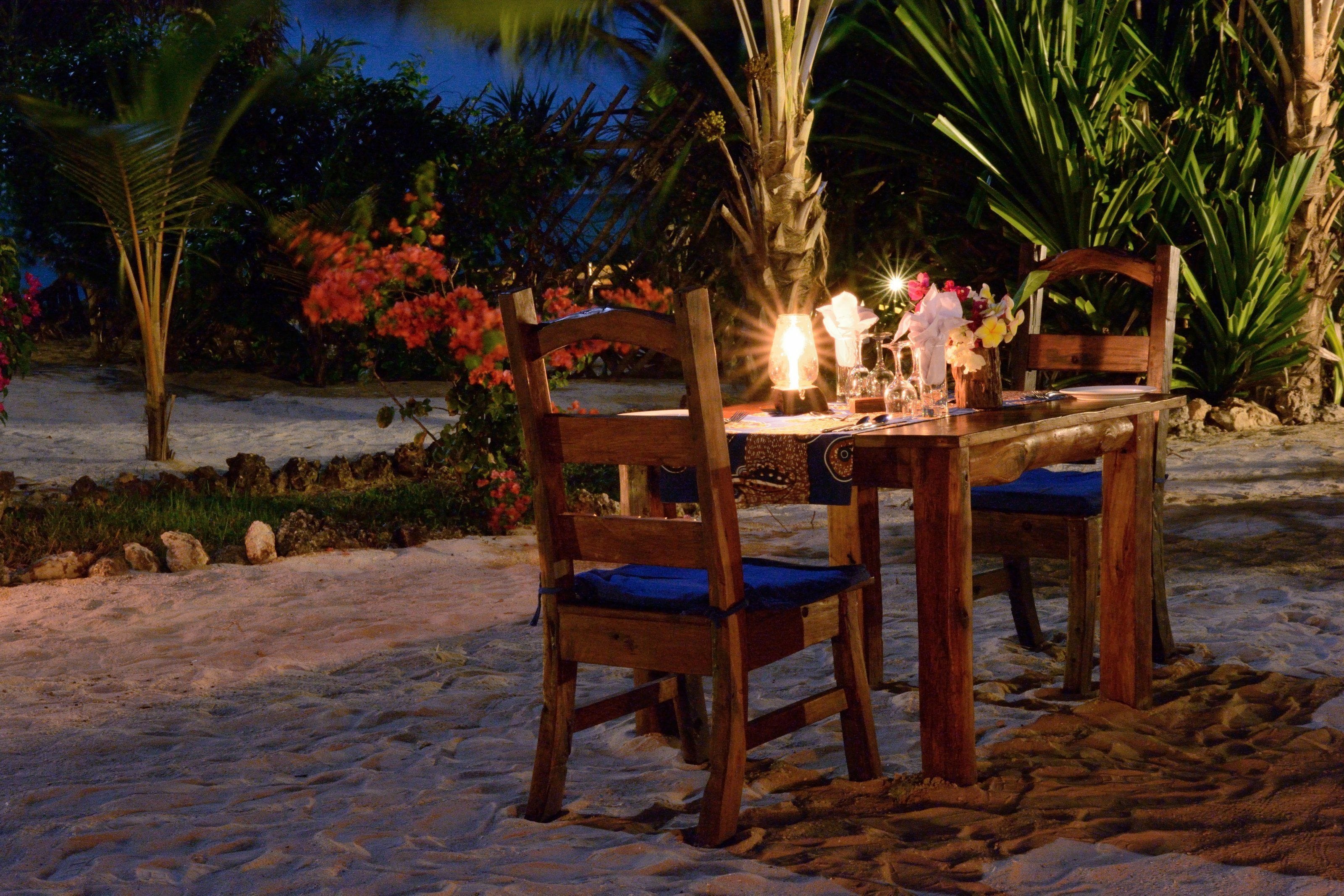 A wooden table with chairs set on a sandy beach at night, surrounded by tropical plants and flowers.