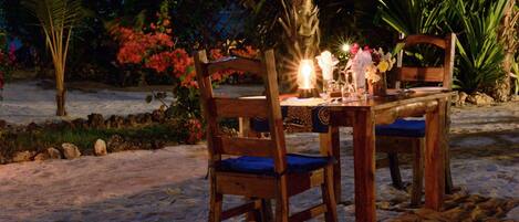 A wooden table with chairs set on a sandy beach at night, surrounded by tropical plants and flowers.