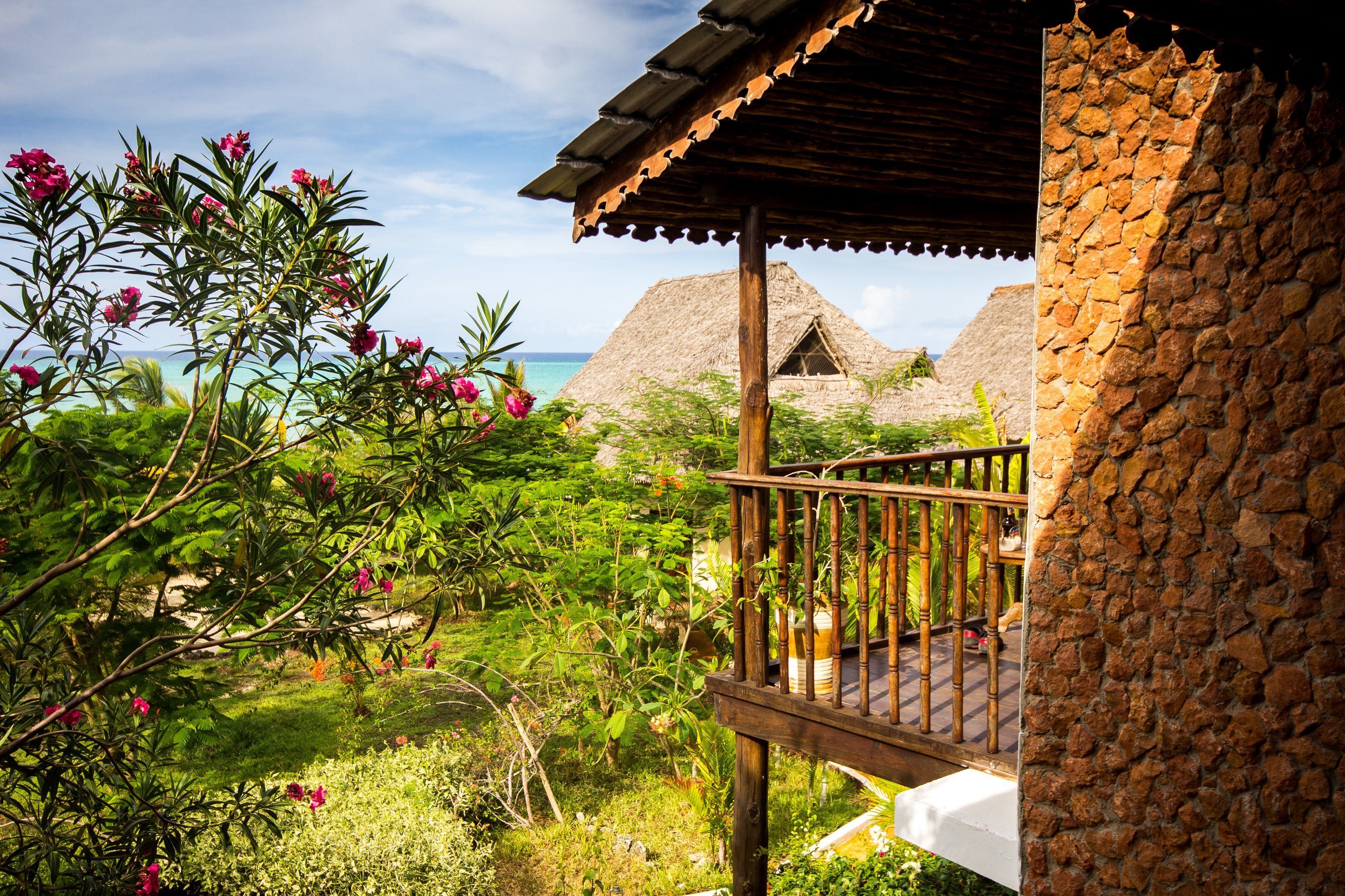 A wooden balcony with a stone wall, overlooking a lush garden and a thatched-roof building by the sea.