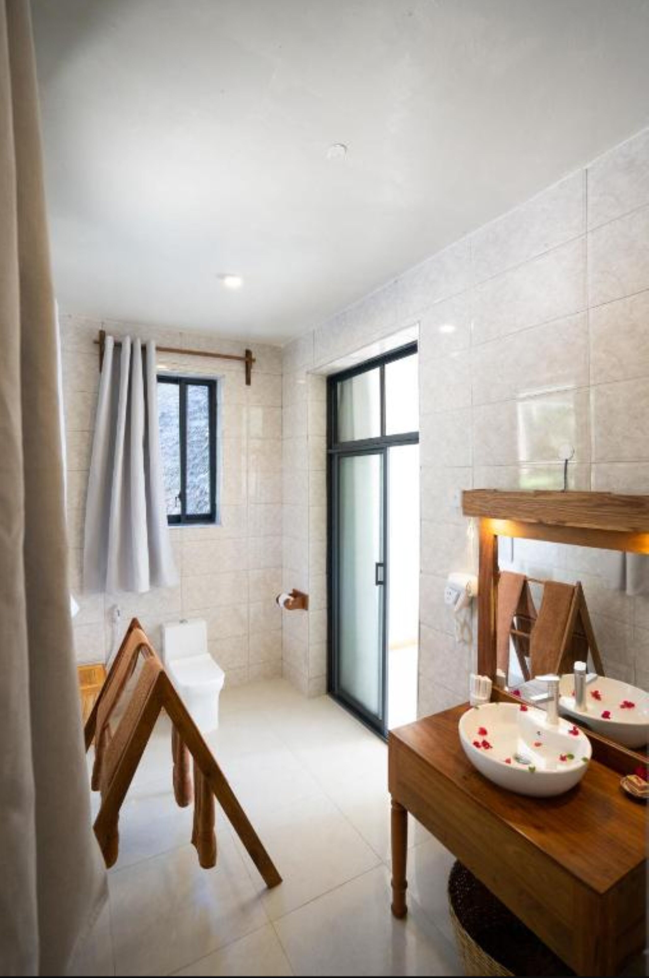 Une salle de bain moderne dotée d’une grande fenêtre, d’un banc en bois, de toilettes blanches et d’un meuble-lavabo en bois avec un vasque.