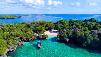 An aerial view of a tropical island with clear turquoise waters, dense green vegetation, and a small boat anchored near a sandy beach.