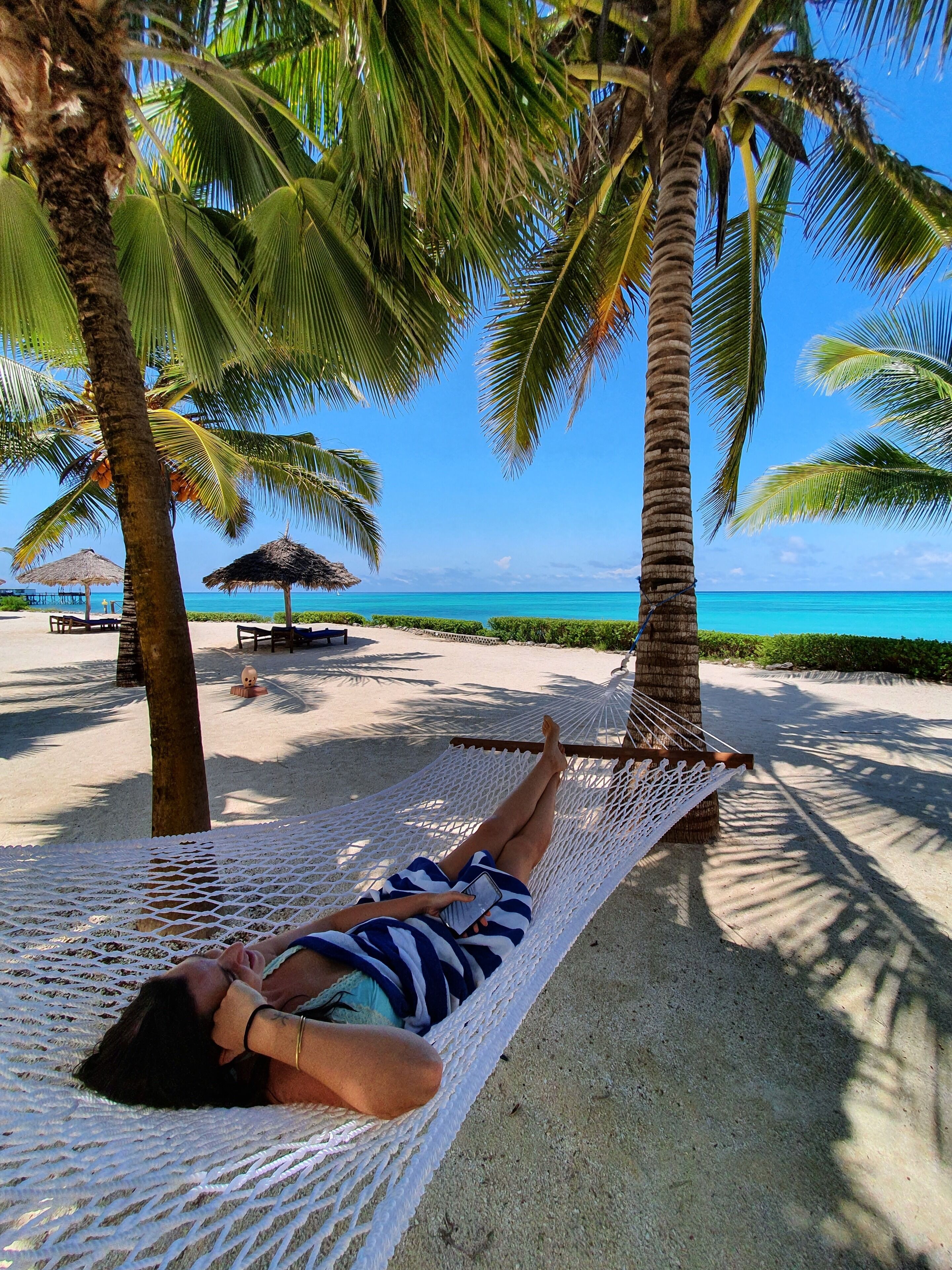 Une personne allongée dans un hamac entre deux palmiers, sur une plage de sable, avec un ciel bleu dégagé et l’océan en arrière-plan.