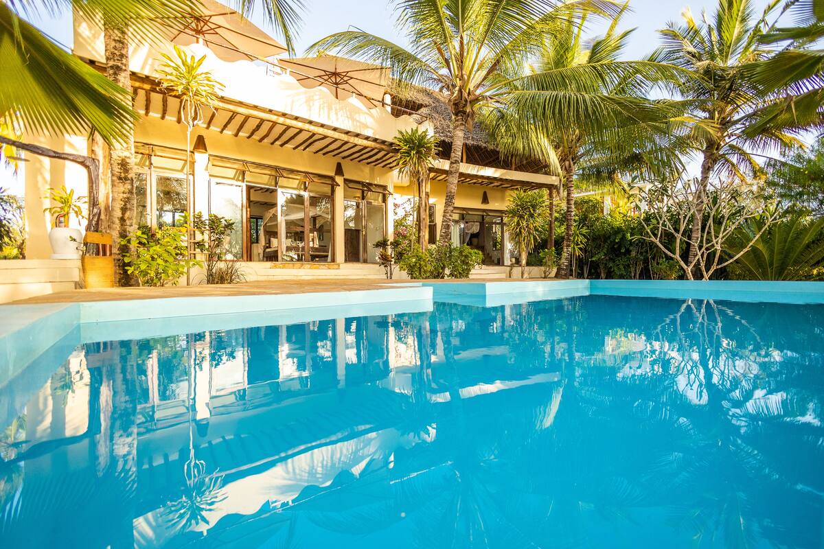 A swimming pool with clear blue water, surrounded by palm trees and a building with large windows and a covered patio area.