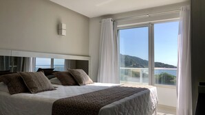 Un dormitorio con una cama grande, un cabecero, una ventana con cortinas y vista al mar.