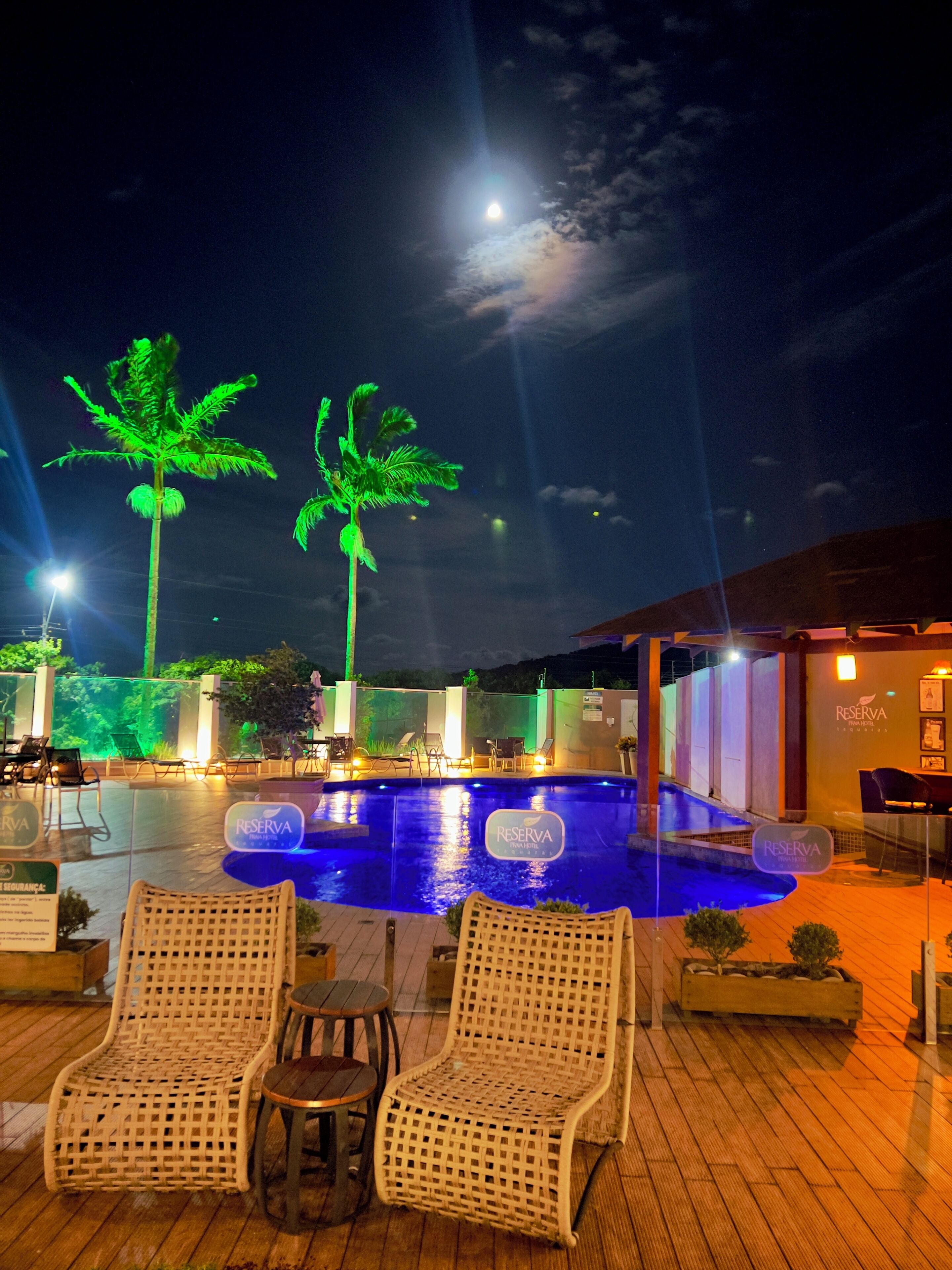 Area piscina illuminata di notte, con poltrone relax e un cartello con la scritta 'RESERVA'.
