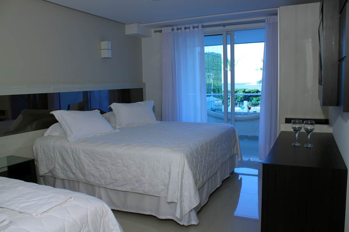 A hotel room with a large bed, a smaller bed, a desk with two wine glasses, and a view of a balcony with outdoor furniture.
