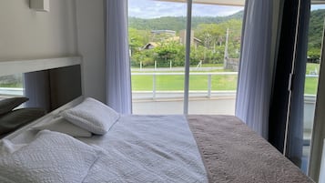 Un dormitorio con un ventanal grande, una cama con ropa de cama blanca y vistas a un paisaje verde y edificios.
