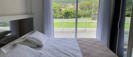 Un dormitorio con un ventanal grande, una cama con ropa de cama blanca y vistas a un paisaje verde y edificios.