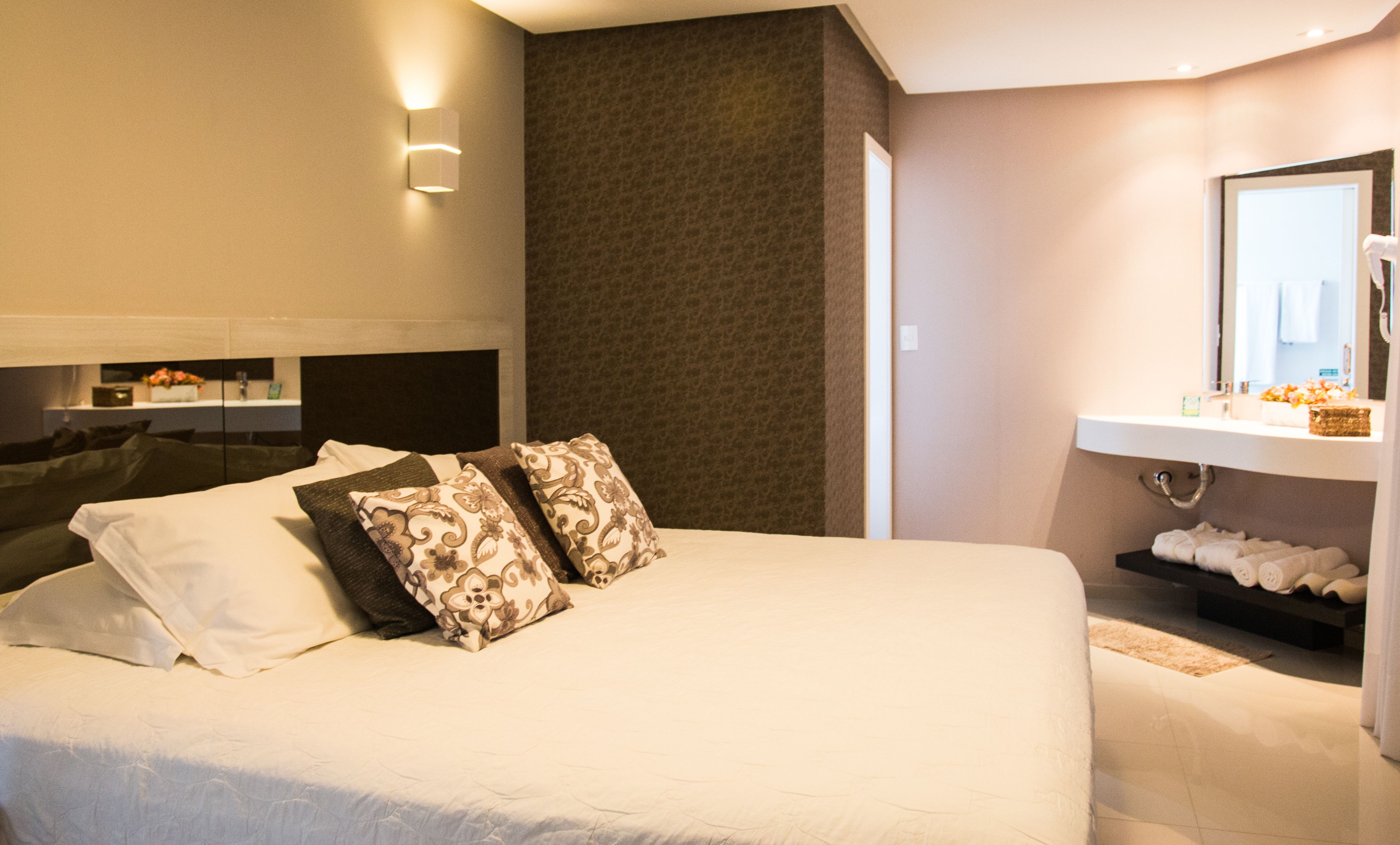 Habitación de hotel con una cama grande, una mesita de noche, un espejo y un baño con lavabo y toallero.