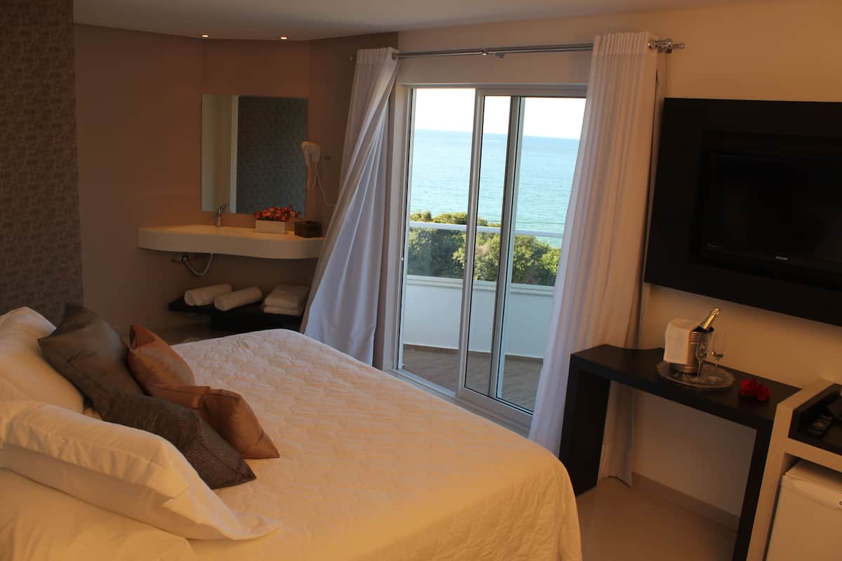 A hotel room with a bed, a bathroom with a sink, a TV, and a balcony with a view of the sea.