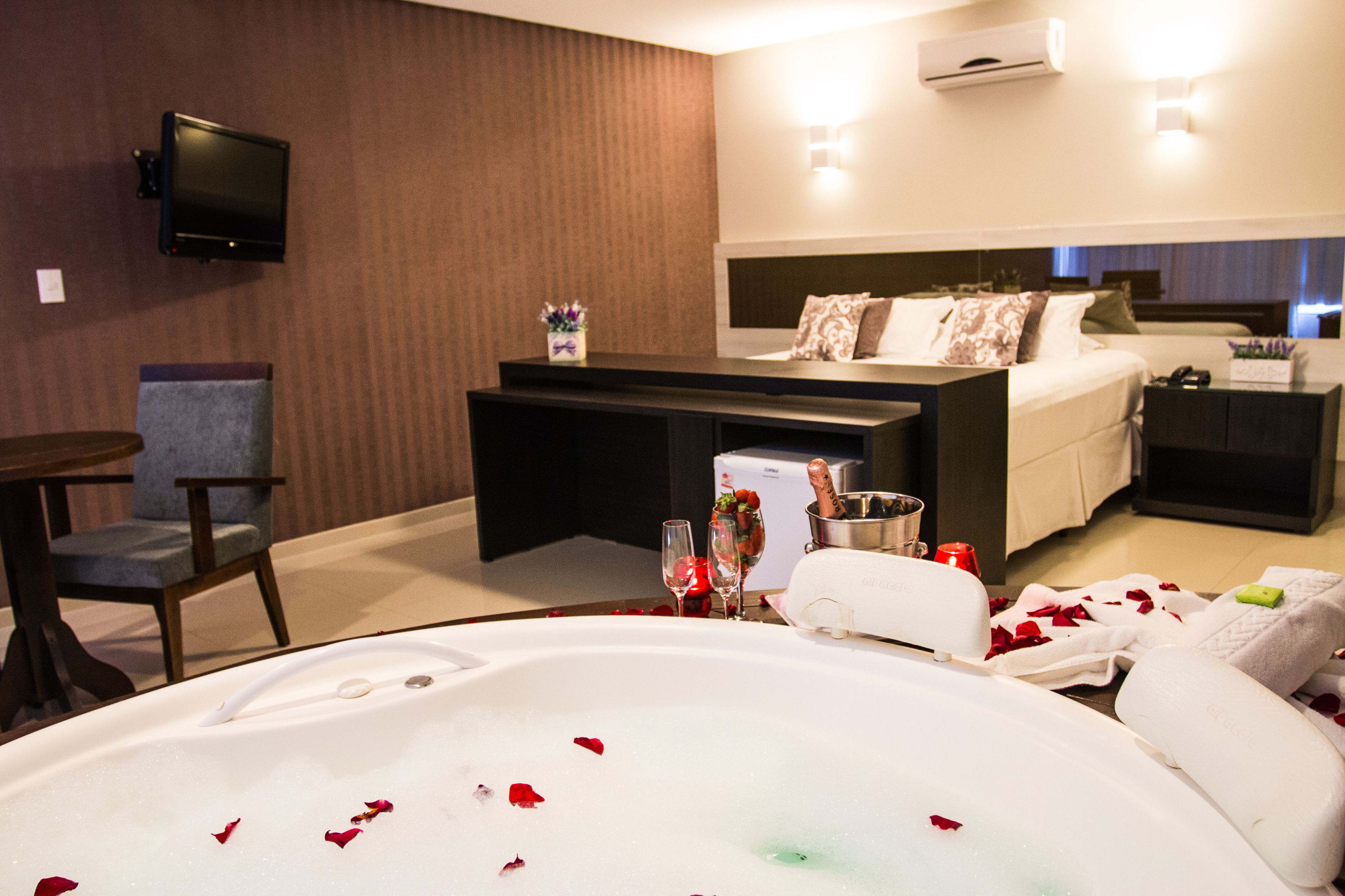A hotel room with a bed, a desk, a chair, a TV, and a bathtub with rose petals.