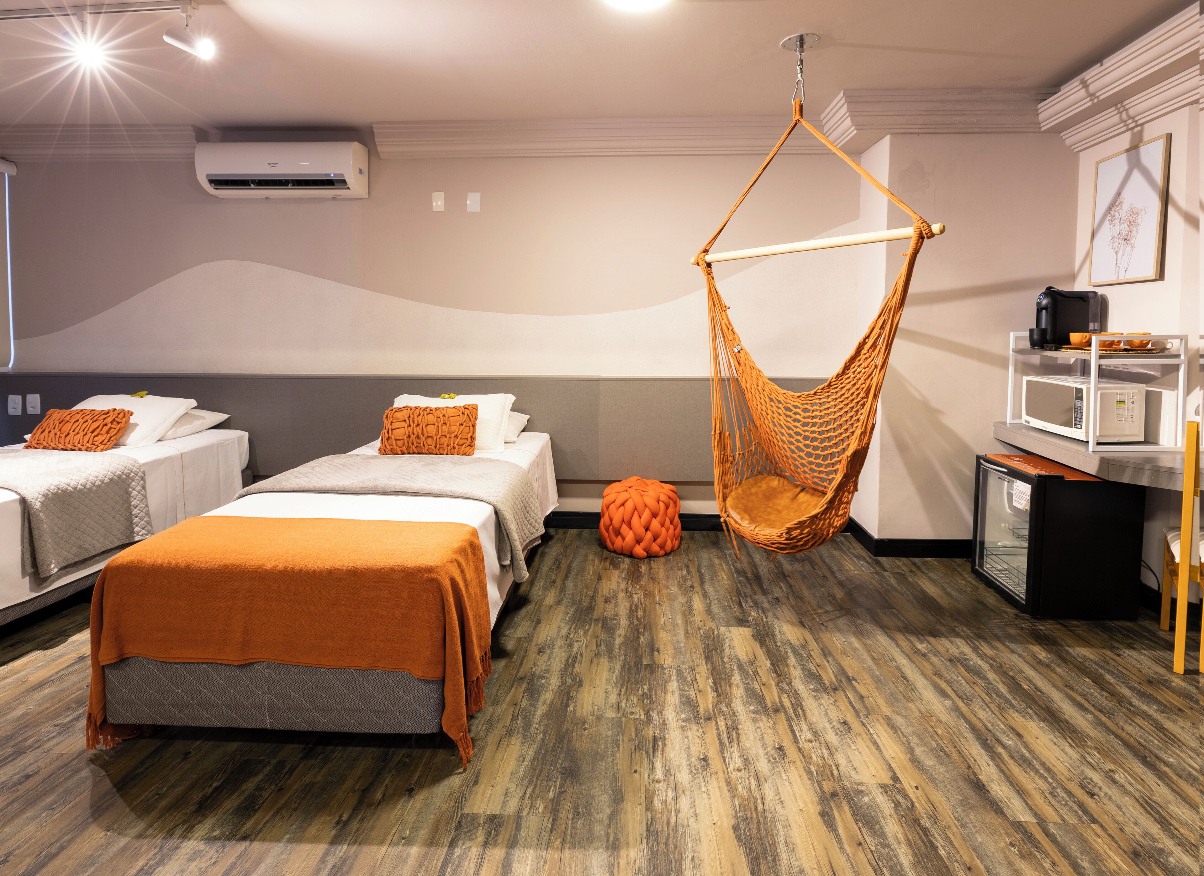 Quarto de hotel com duas camas, uma cadeira suspensa, um banquinho redondo, um micro-ondas e um frigorífico.