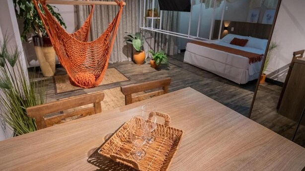 Une chambre moderne avec une table à manger en bois, un hamac orange suspendu, un lit avec une tête de lit blanche et un téléviseur fixé à un mur blanc.