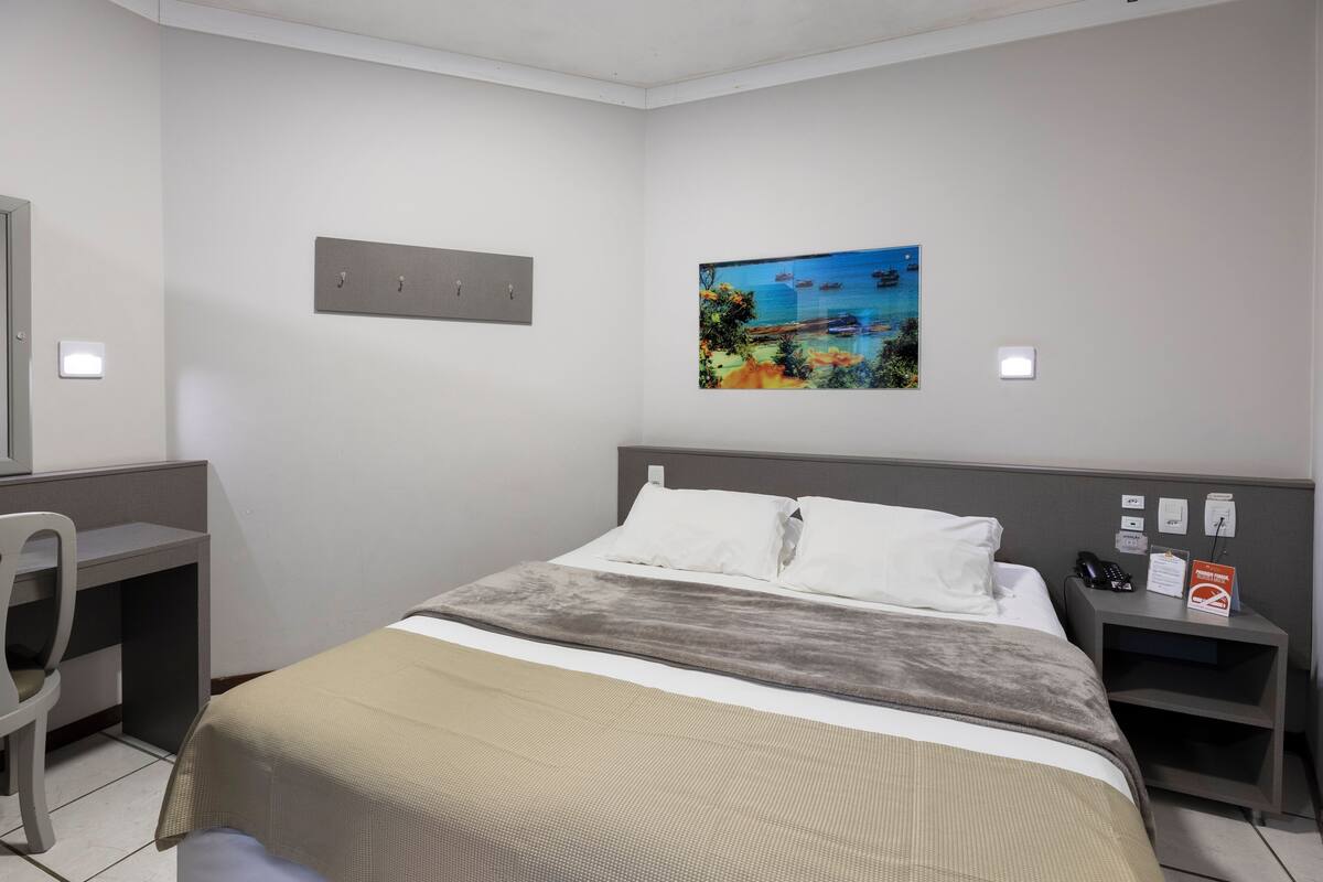 A hotel room with a bed, a desk, a chair, a nightstand, and a painting on the wall.