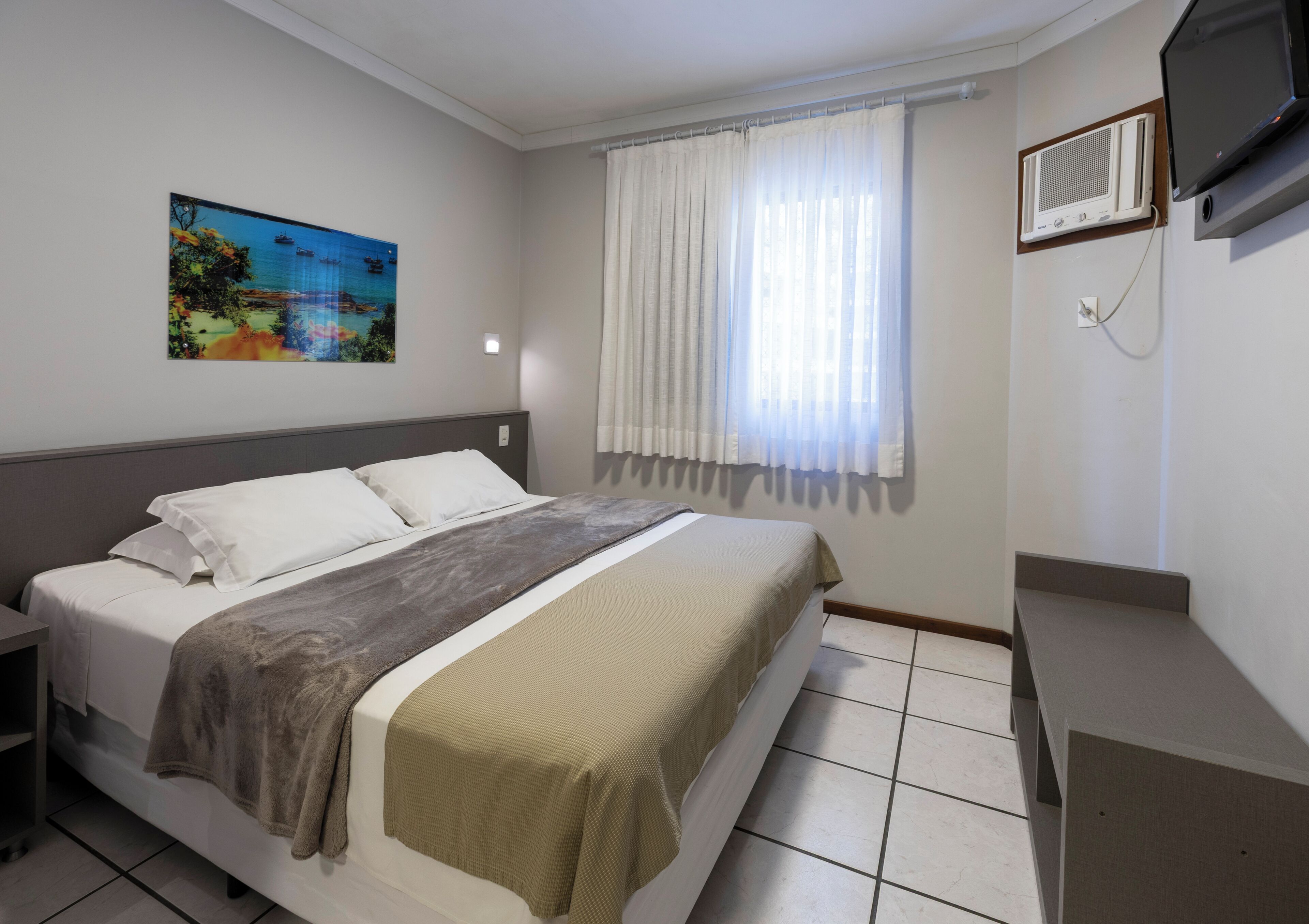 Una habitación de hotel con cama, un cuadro, una ventana con cortinas, televisor y aire acondicionado.