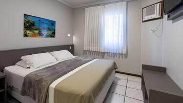 Una habitación de hotel con cama, un cuadro, una ventana con cortinas, televisor y aire acondicionado.