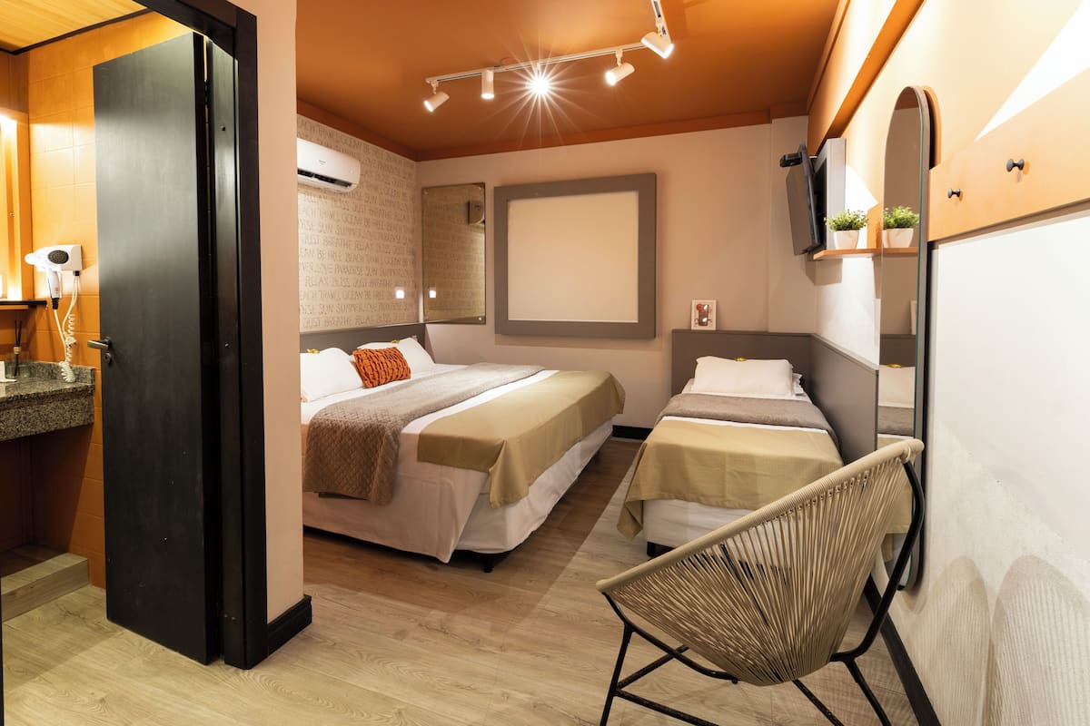 A hotel room with two beds, a chair, a mirror, and a small shelf with plants.