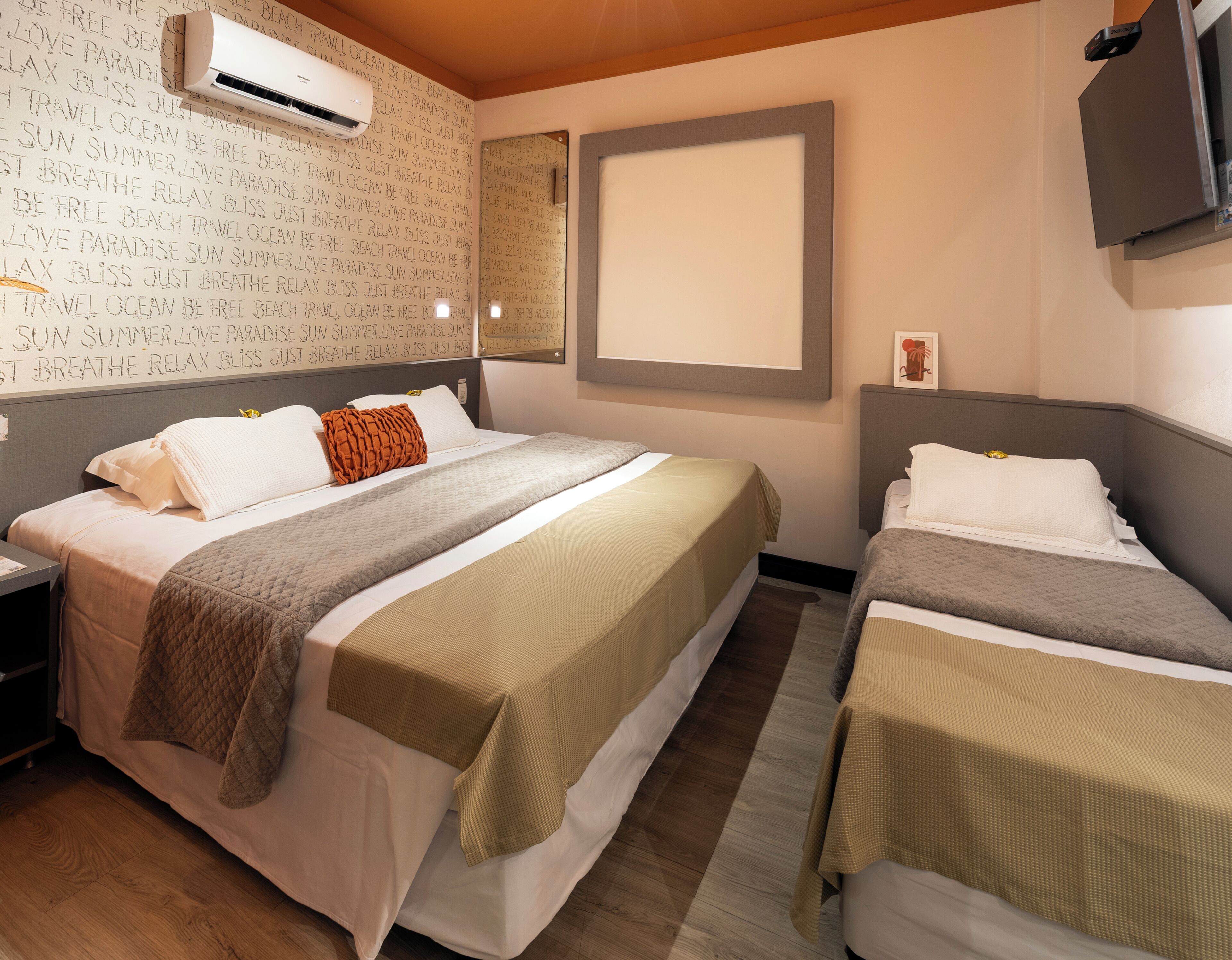 Habitación de hotel con dos camas, un equipo de aire acondicionado grande, un televisor de pantalla plana y una pared con texto relacionado con viajes.