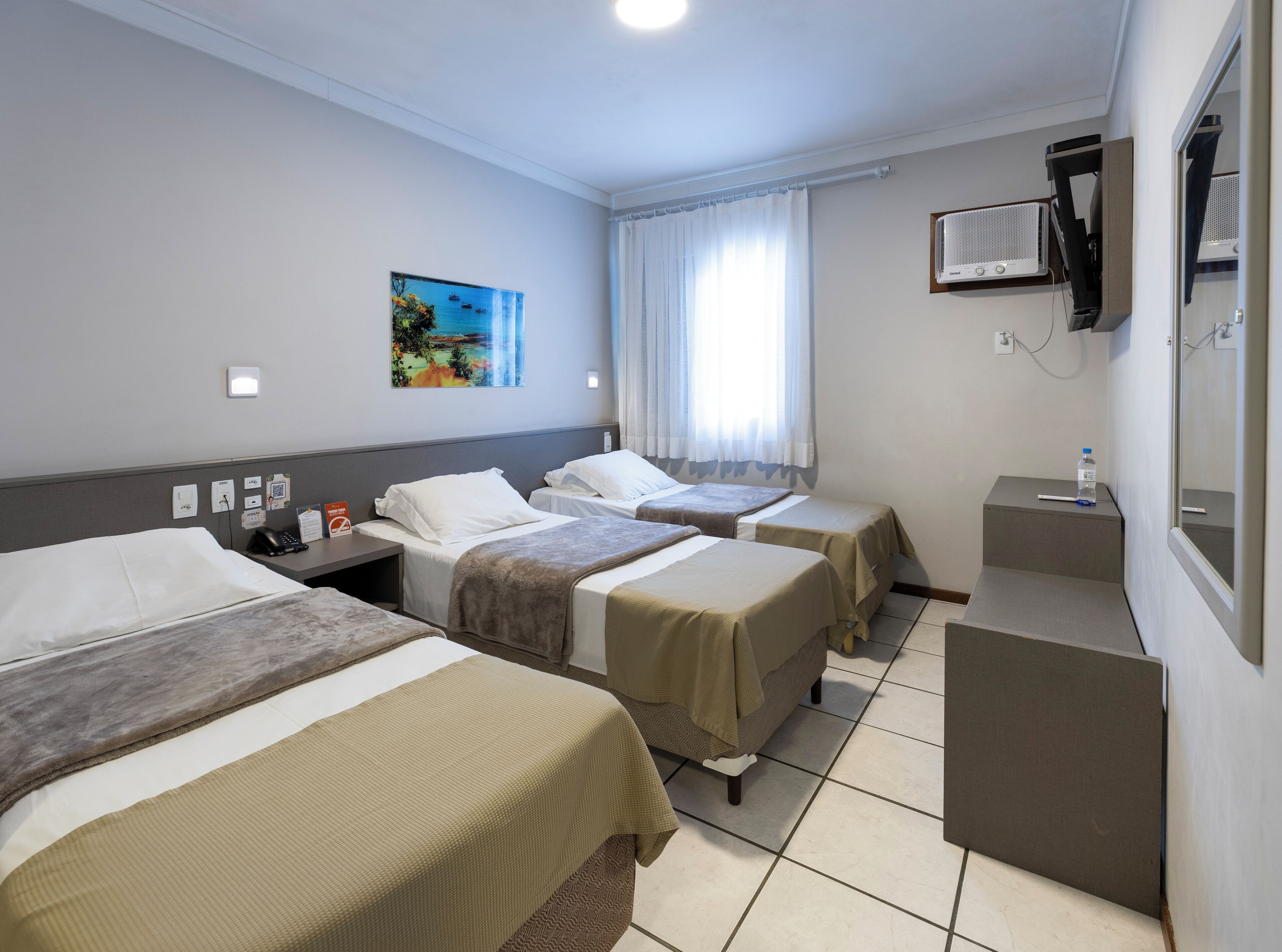 A hotel room with three beds, a desk, a chair, a painting, and a window with curtains.