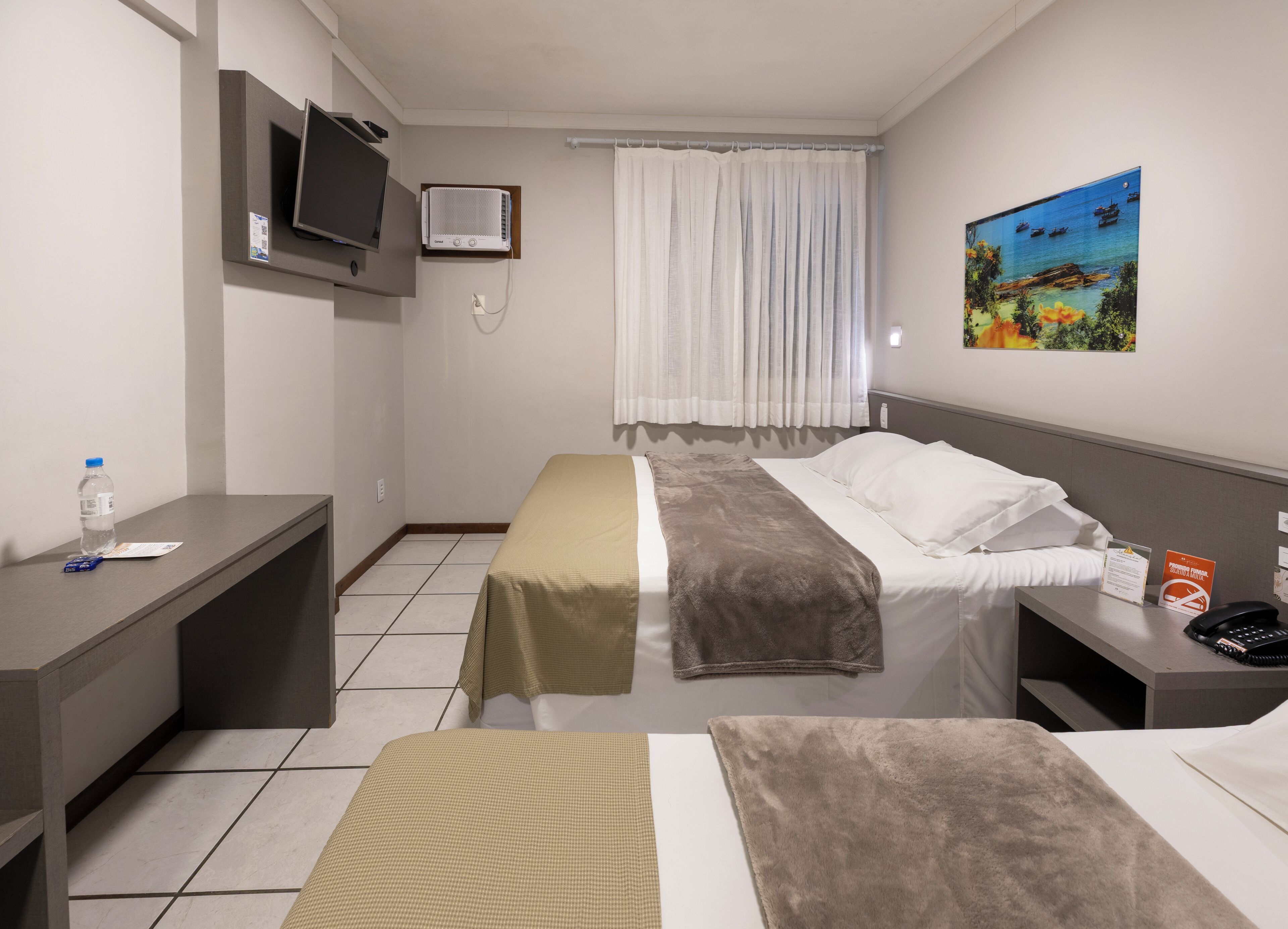 A hotel room with a bed, bedside table, TV, air conditioning unit, window with curtains, and a painting on the wall.