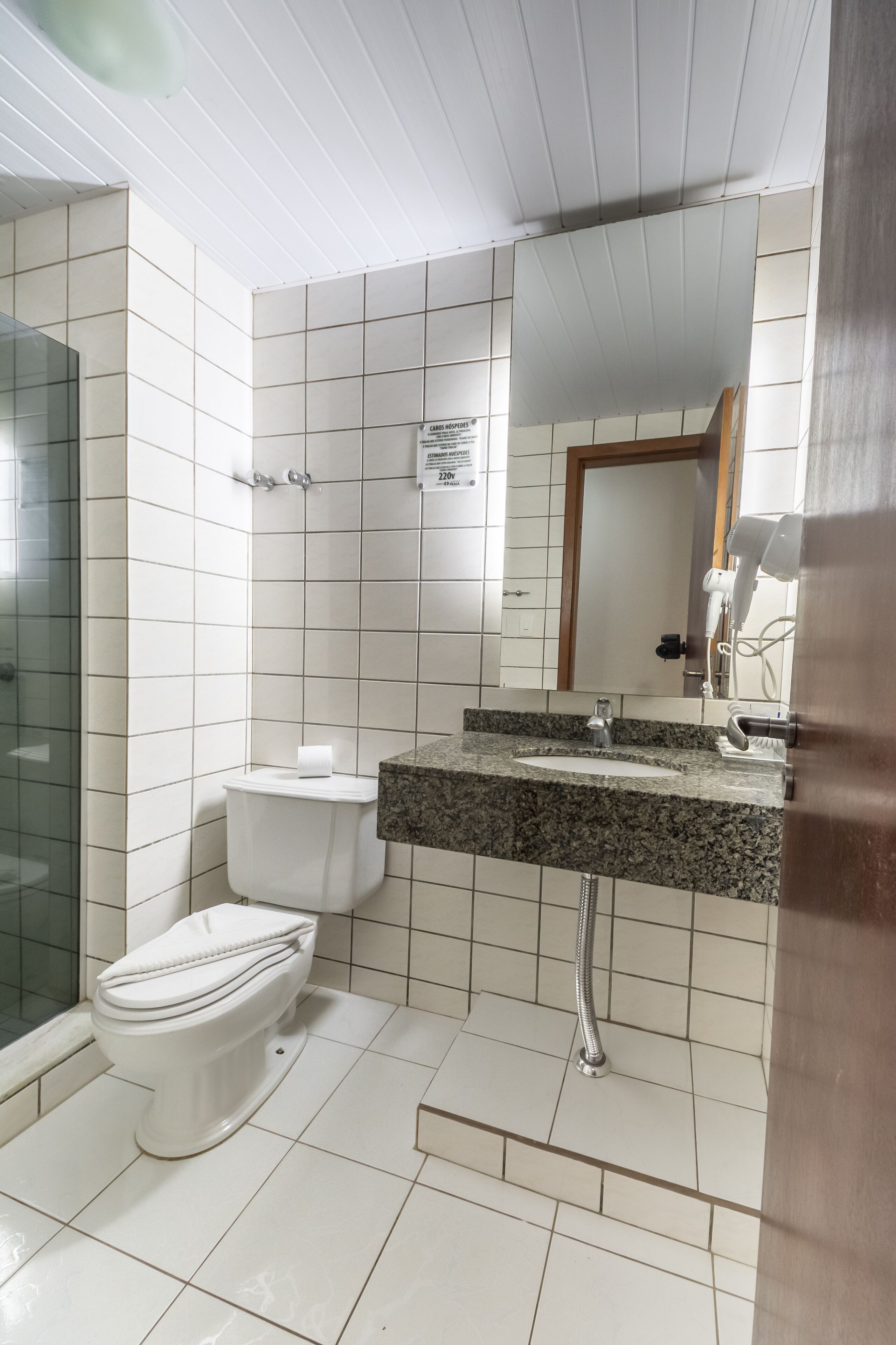 A bathroom with white tiled walls, a white toilet, a granite countertop sink, and a shower with a glass enclosure.