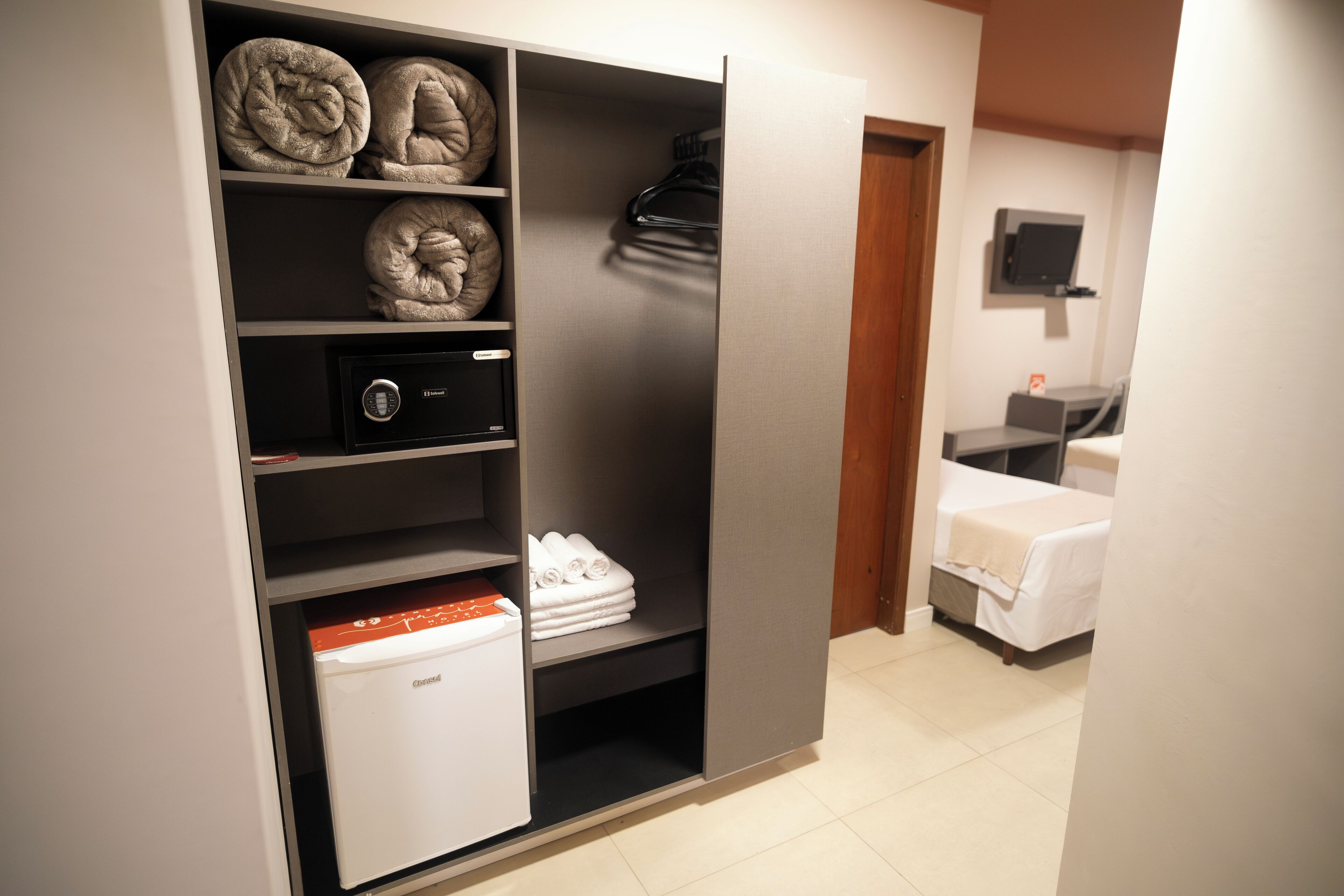 Habitación de hotel con minibar, caja fuerte y toallas ordenadamente dobladas en estantes.