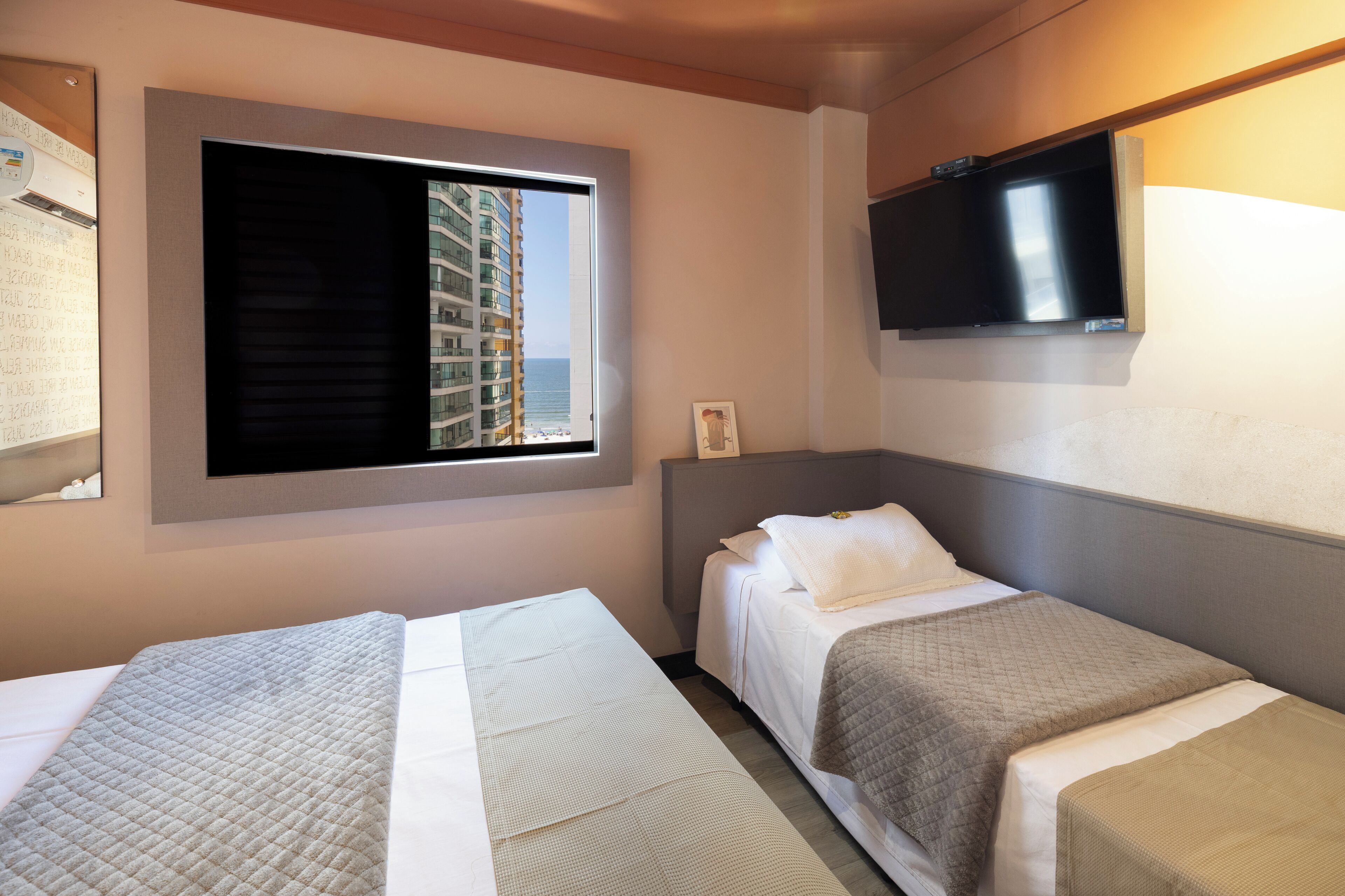 Ein Hotelzimmer mit zwei Betten, einem an der Wand montierten Fernseher und einem Fenster mit Blick auf ein Gebäude.