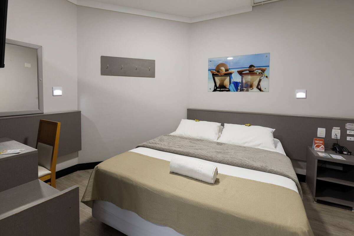 A hotel room with a bed, a desk, a chair, and a painting on the wall.