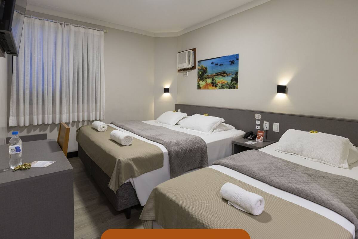 Ein Hotelzimmer mit zwei Betten, einem Schreibtisch, einem Stuhl, einer Klimaanlage, einem Gemälde und einem Fenster mit Vorhängen.