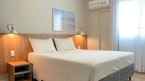 A neatly made bed with a grey headboard, two bedside lamps, and a small wooden nightstand. - Hotel Itália (Balneário Camboriú)