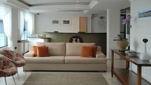 A living room with a beige sofa, orange cushions, a wooden side table, and a patterned rug. - Hotel Itália (Balneário Camboriú)