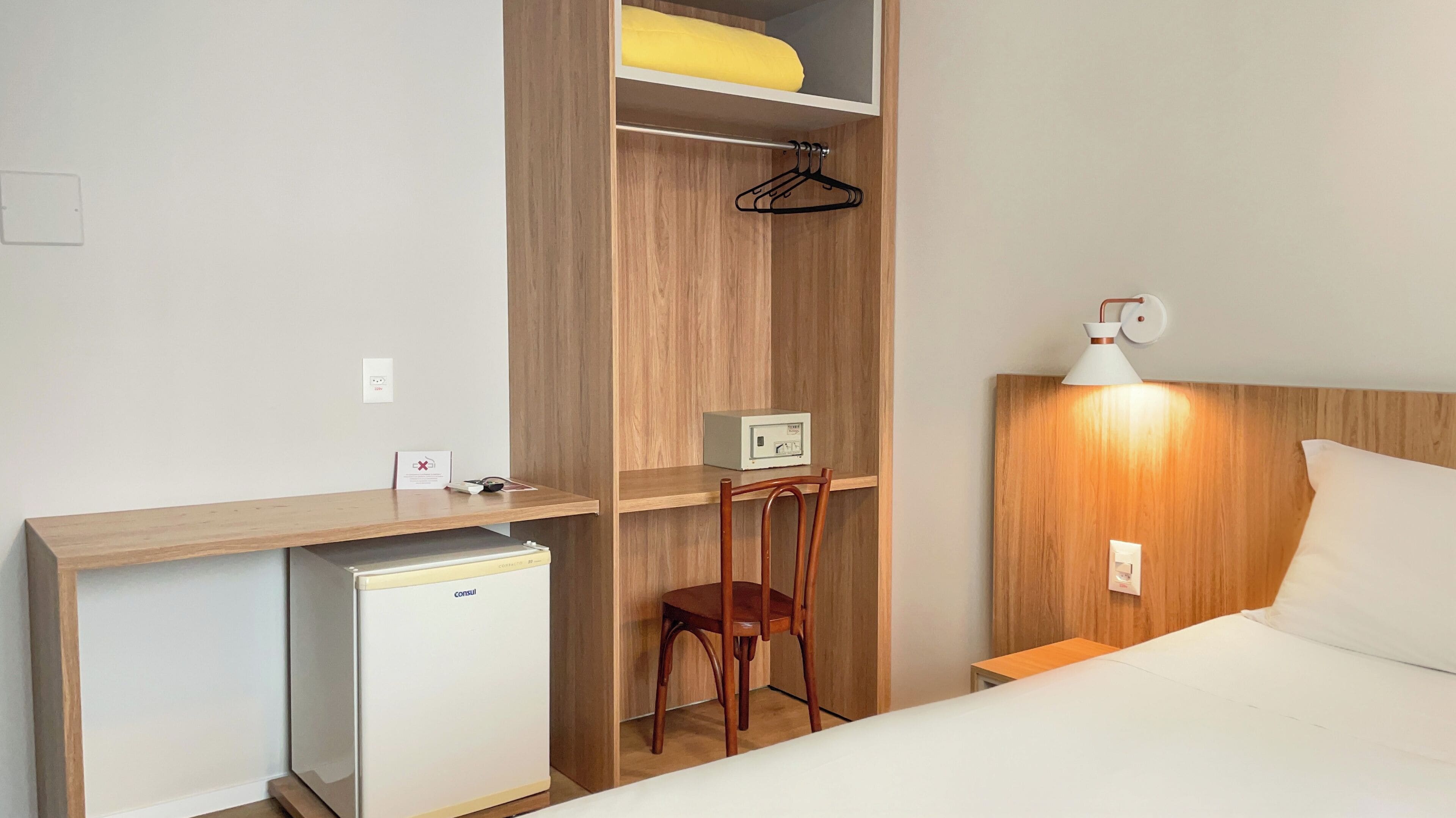 A hotel room with a bed, bedside table, lamp, and a small desk with a chair and a refrigerator.