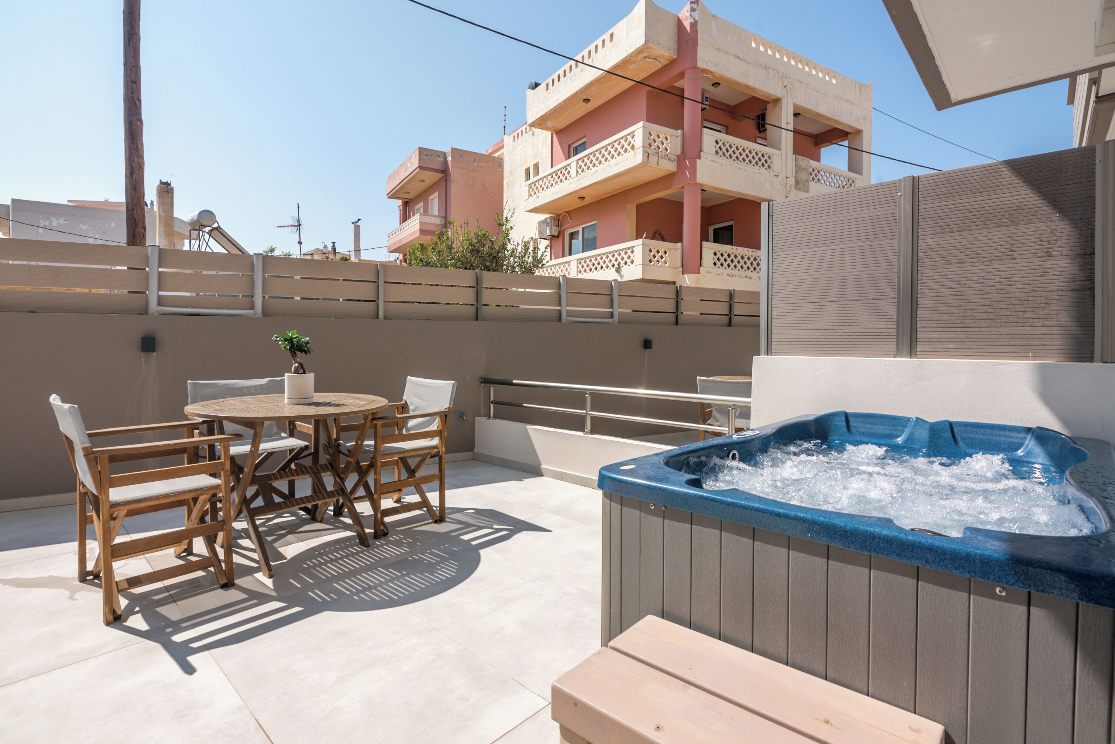 Une terrasse extérieure dotée d’un jacuzzi, de meubles en bois et offrant une vue sur un bâtiment rose en arrière-plan.