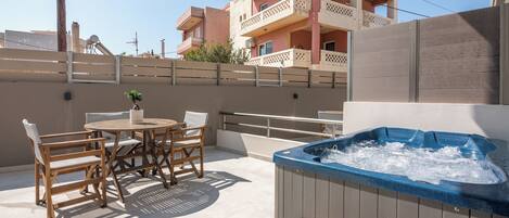 Une terrasse extérieure dotée d’un jacuzzi, de meubles en bois et offrant une vue sur un bâtiment rose en arrière-plan.