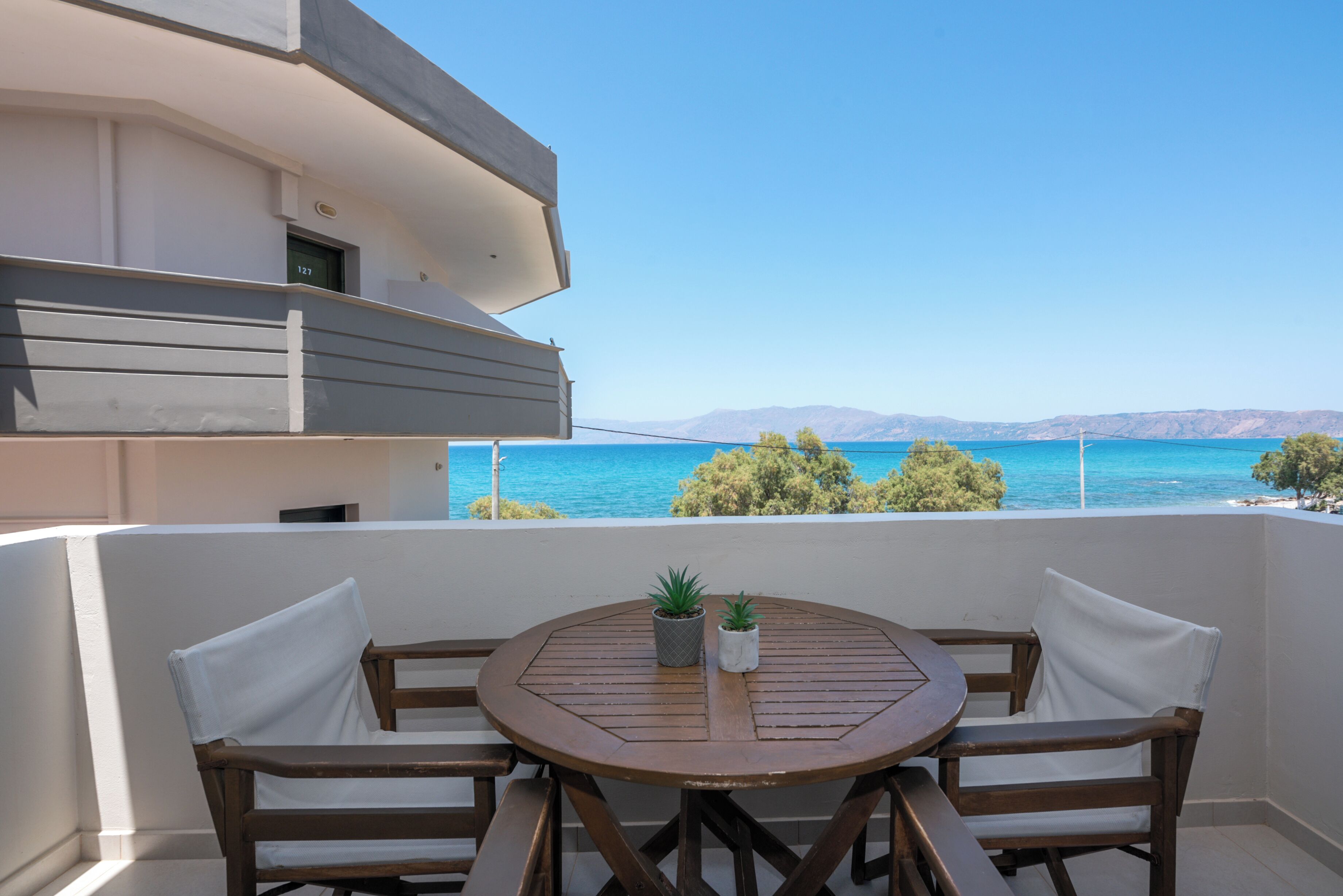 A balcony with a wooden table and two chairs, overlooking a body of water and distant mountains.
