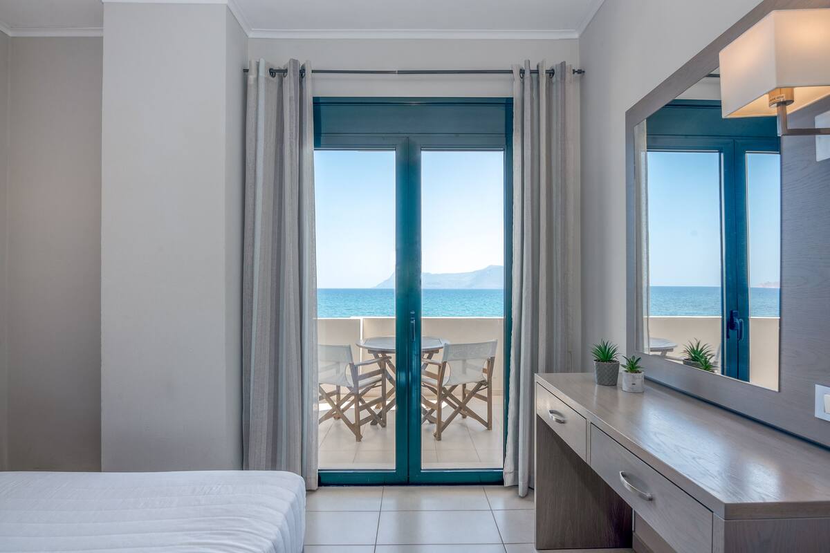 A bedroom with a balcony, a bed, a desk, and a view of the sea.