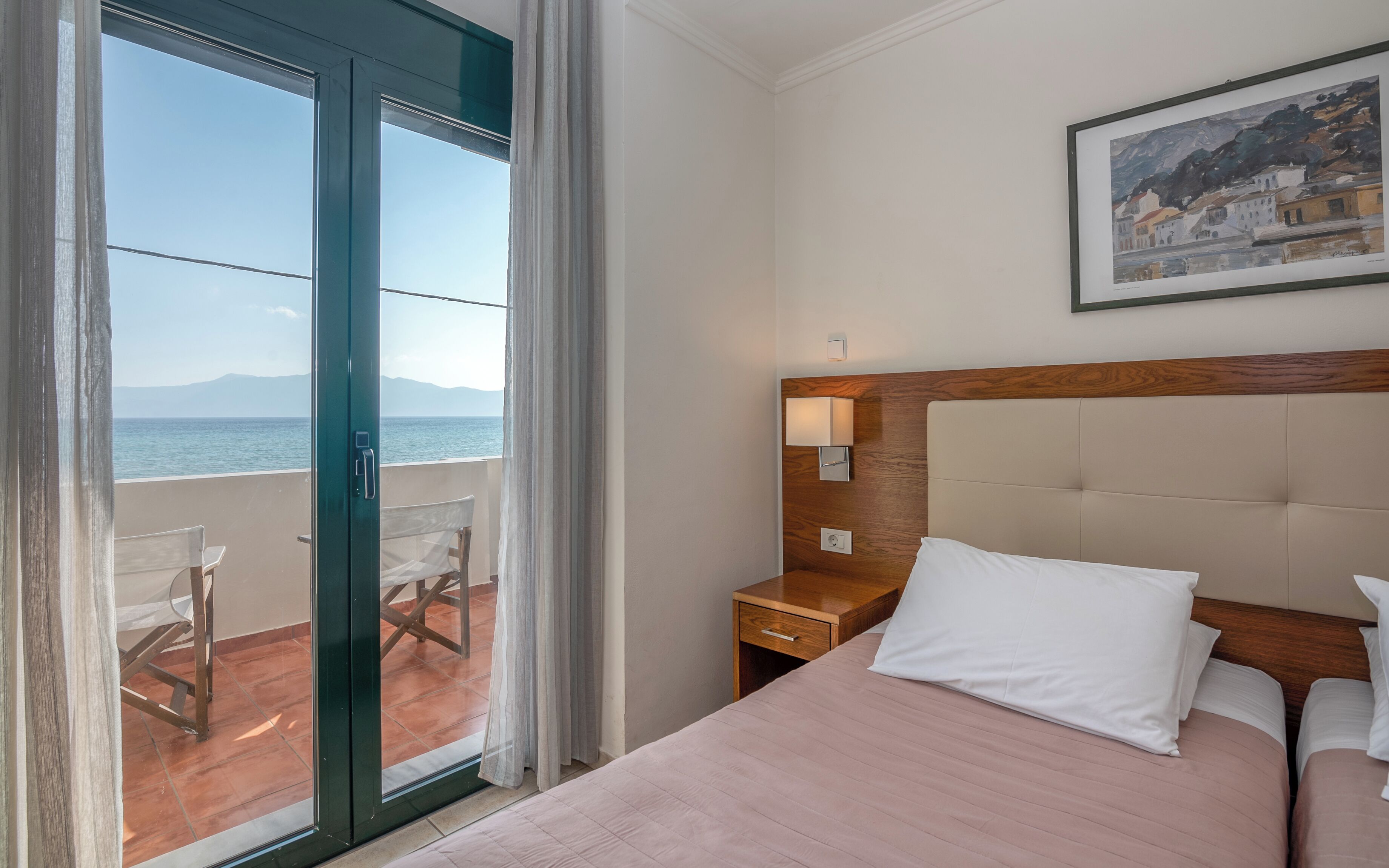 A hotel room with a bed, a nightstand, a lamp, a painting, and a view of the sea through a sliding door.