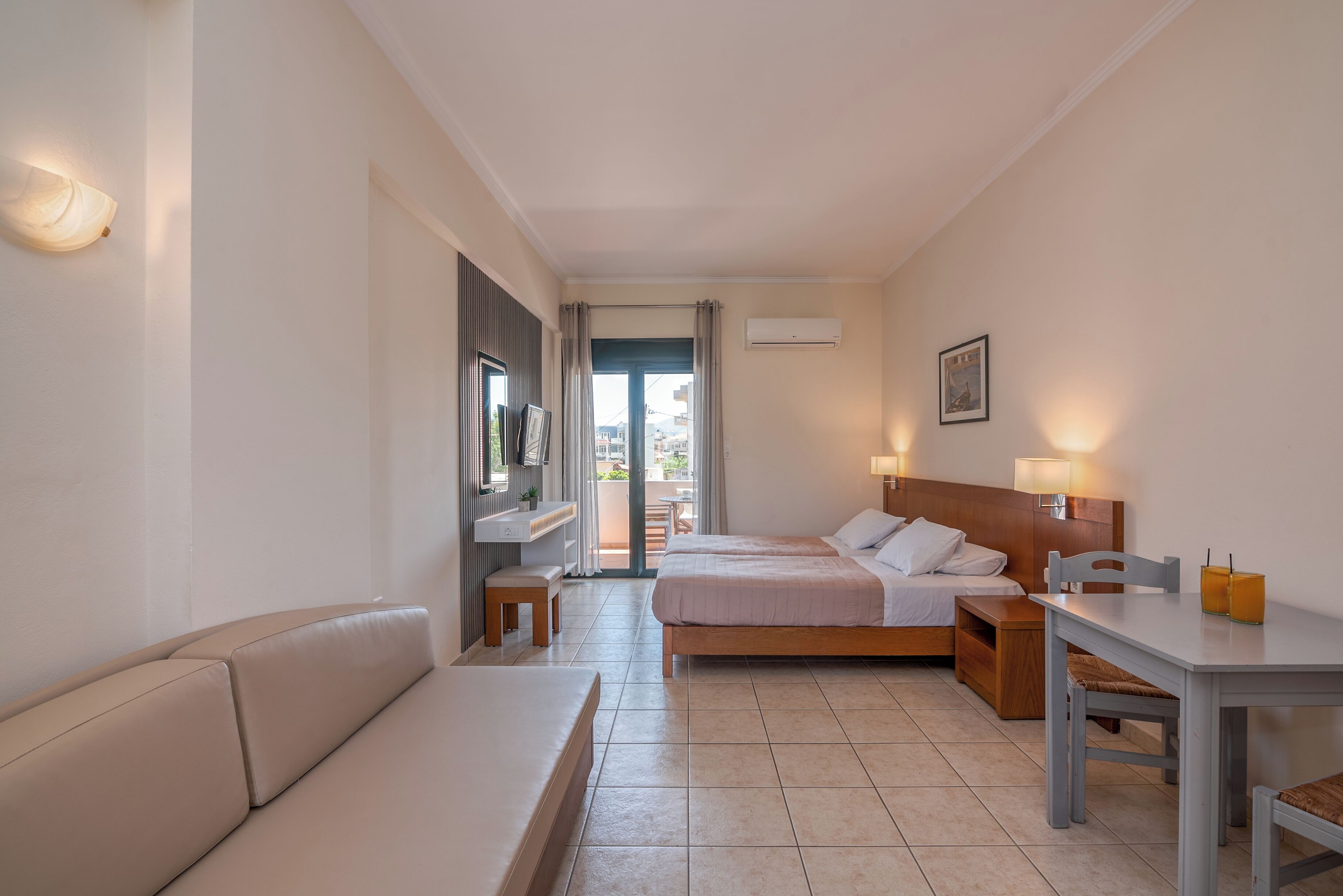 A hotel room with a sofa, two single beds, a table with chairs, a TV, and a balcony with a view.
