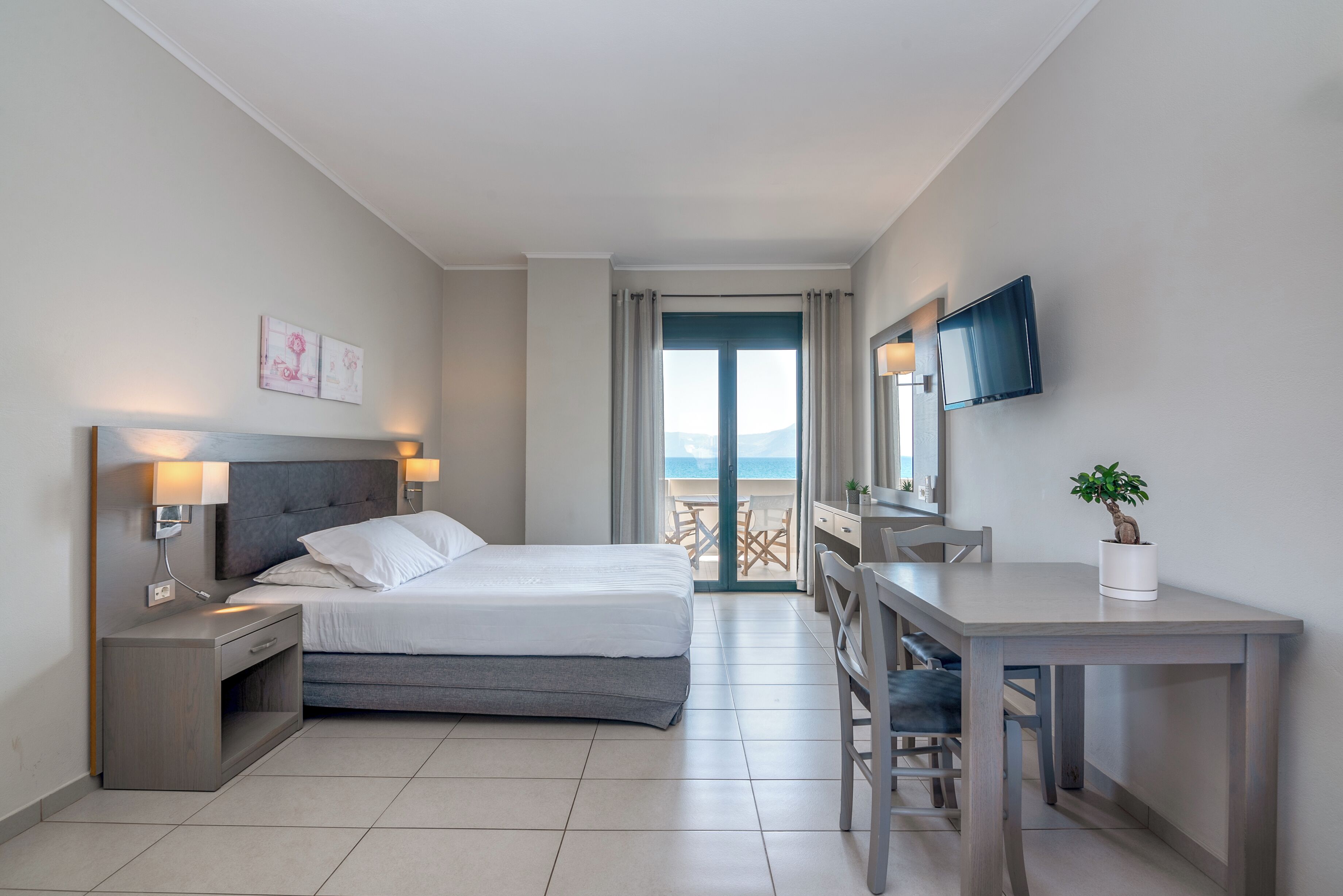 A hotel room with a bed, bedside tables, a desk, a chair, a TV, and a balcony with a view.