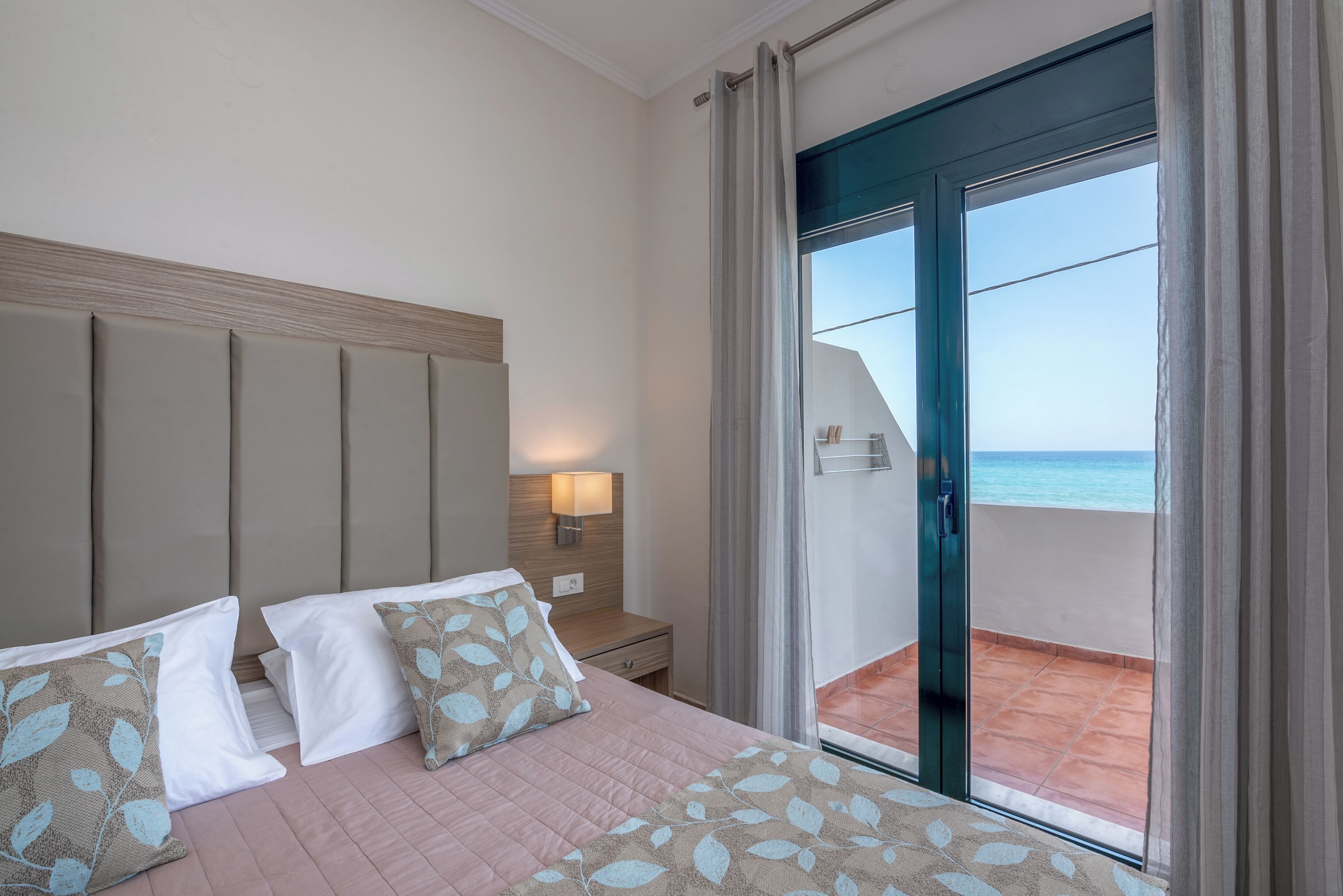 Habitación de hotel con cama, mesita de noche, lámpara y vista al mar a través de una puerta corrediza de vidrio.