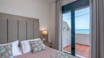 Habitación de hotel con cama, mesita de noche, lámpara y vista al mar a través de una puerta corrediza de vidrio.