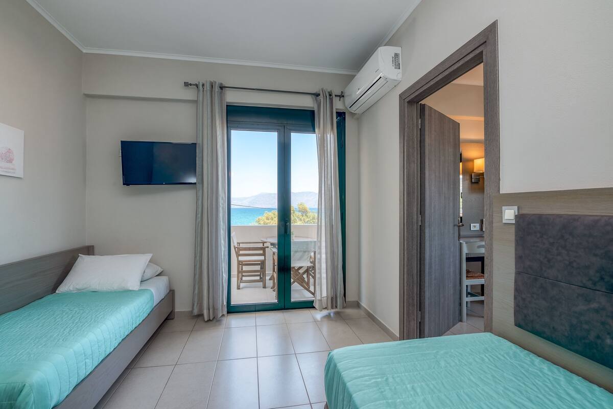 A hotel room with two beds, a TV, an air conditioning unit, and a balcony with a view of the sea.